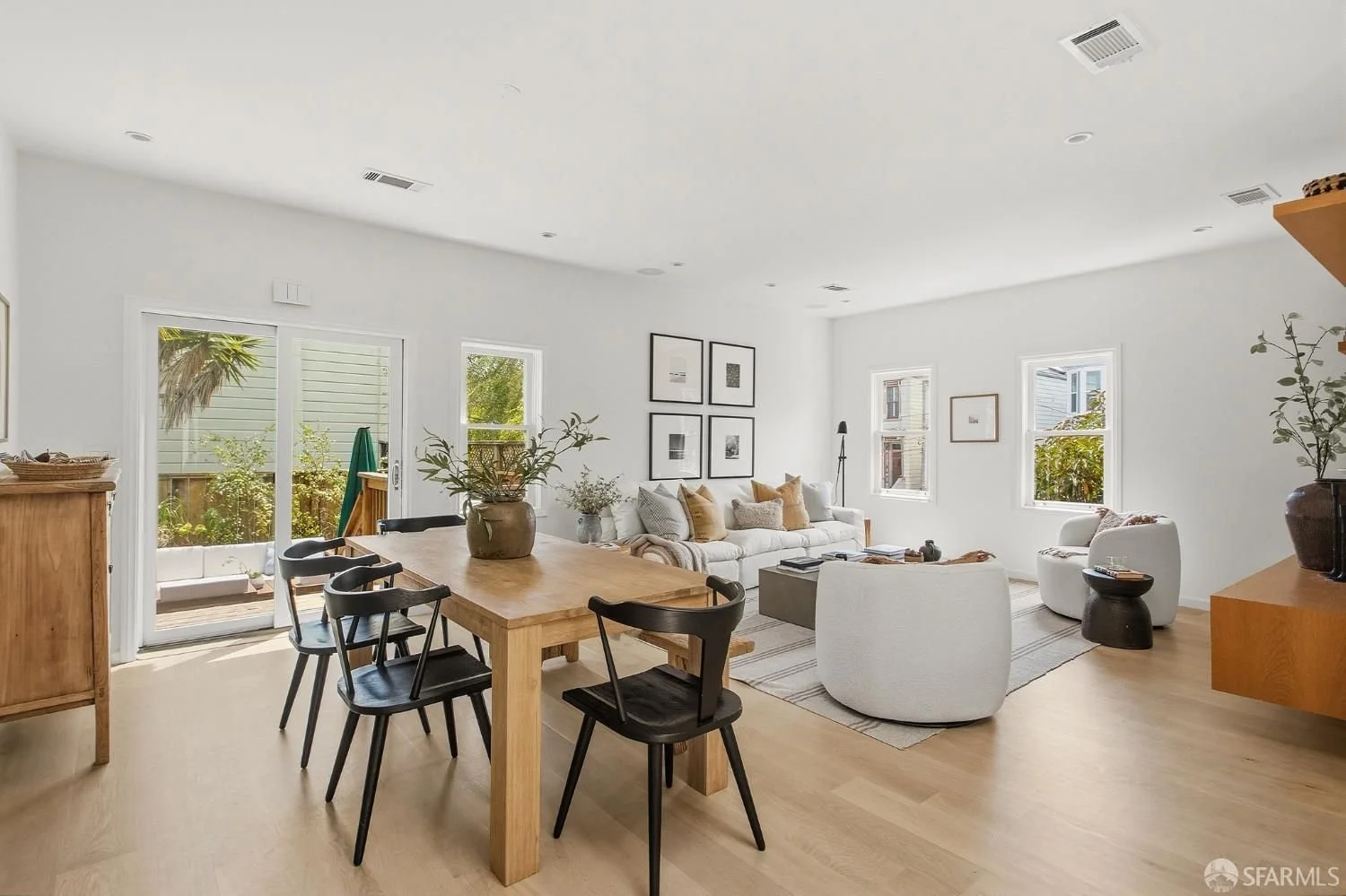 Living room with white walls, a white fireplace, a wooden sideboard, black and white framed photographs, a beige cushioned chair, a vase with branches, and a coffee table with books.