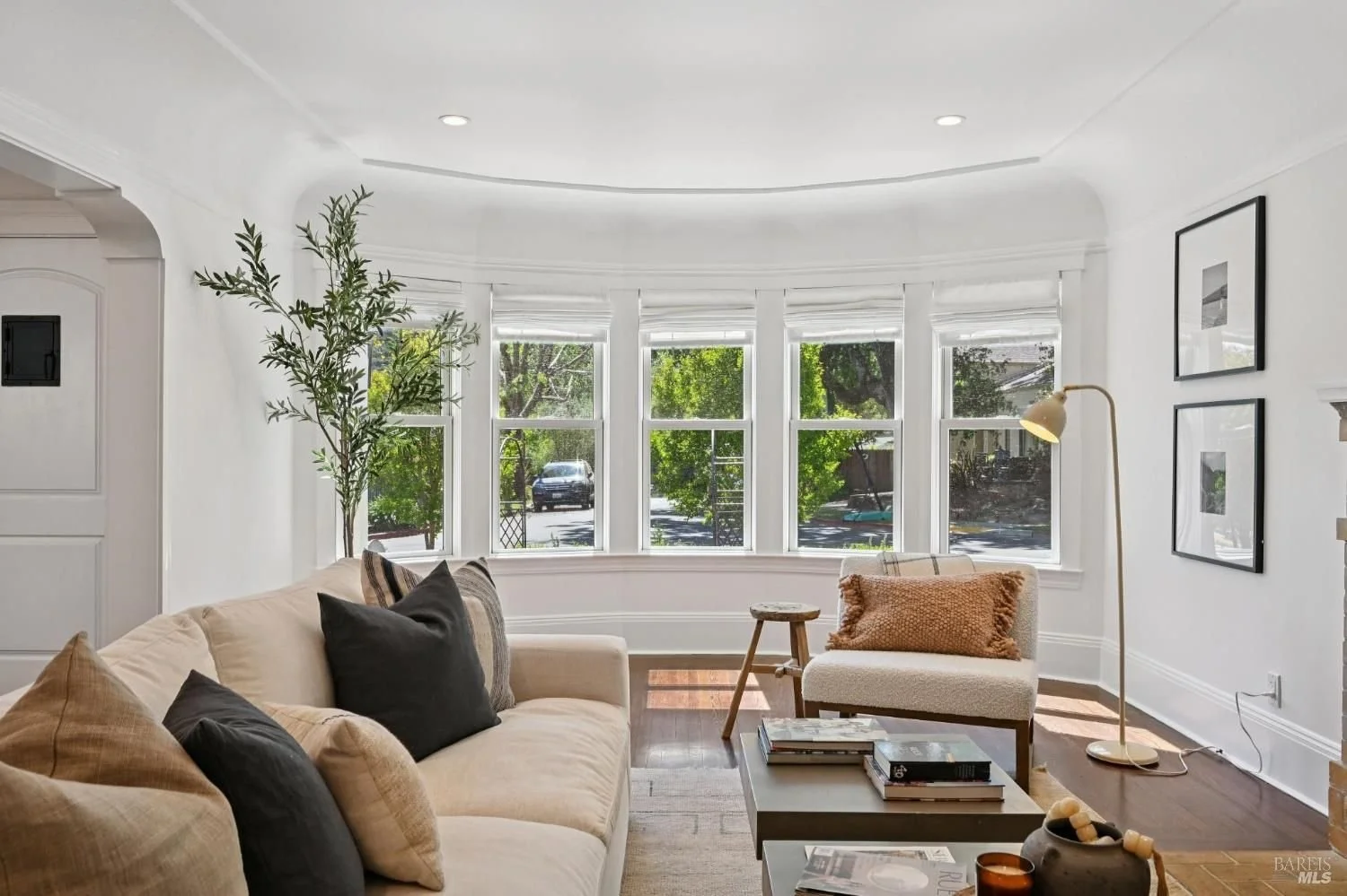 Living room with white walls, a white fireplace, a wooden sideboard, black and white framed photographs, a beige cushioned chair, a vase with branches, and a coffee table with books.