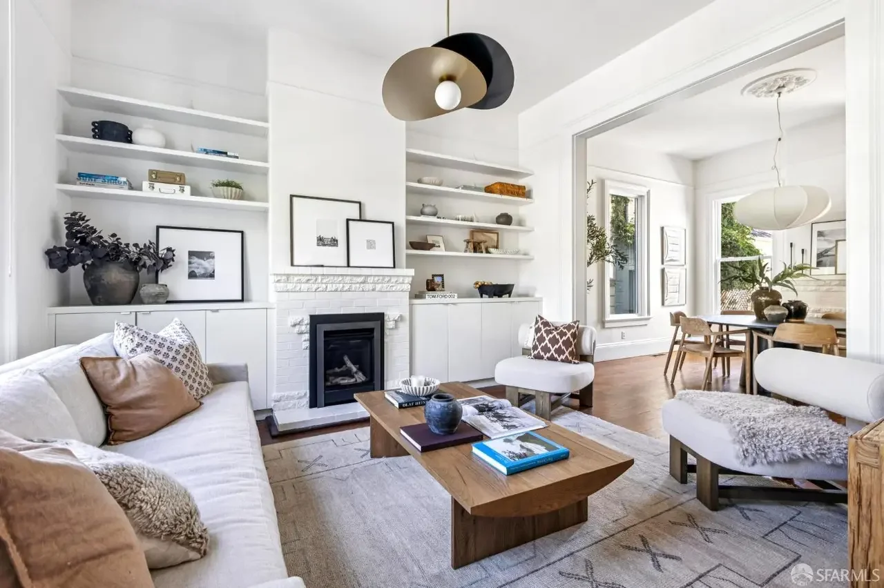 Modern dining area with a white table and six black chairs, open to a kitchen with white upper cabinets, dark lower cabinets, a stainless steel range hood, white subway tile backsplash, and a large potted plant centerpiece on the table.