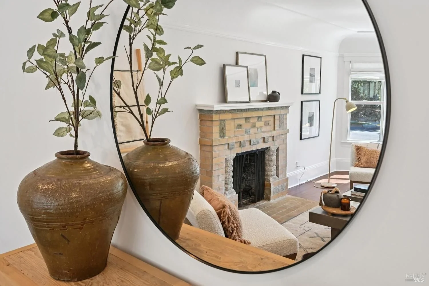 A bright, airy bedroom with two beds, beige headboards, a white dresser, and a large window showing colorful houses outside. Decor includes woven furniture, pillows, and a textured rug.