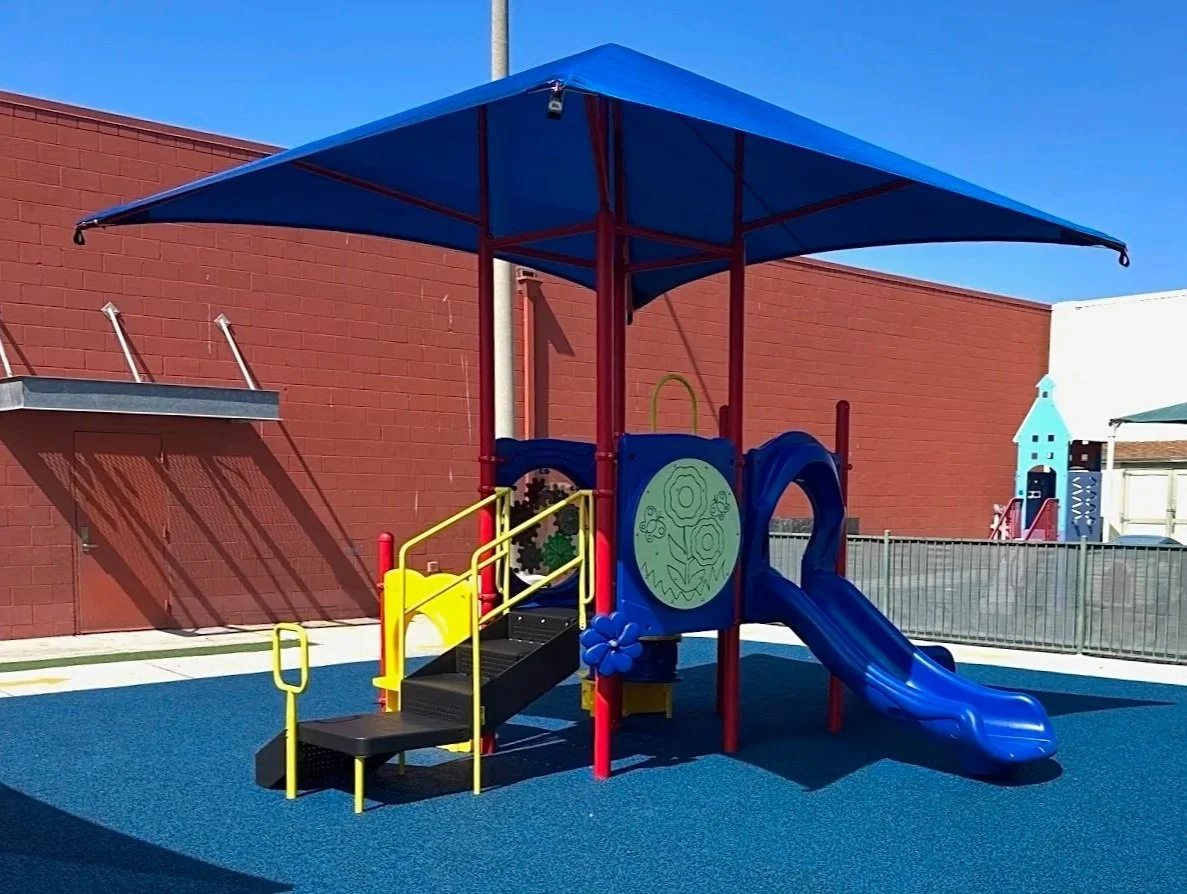 Colorful playground with a blue slide, a small climbing ramp with yellow handrails, and a large shaded blue canopy on a blue rubber surface.