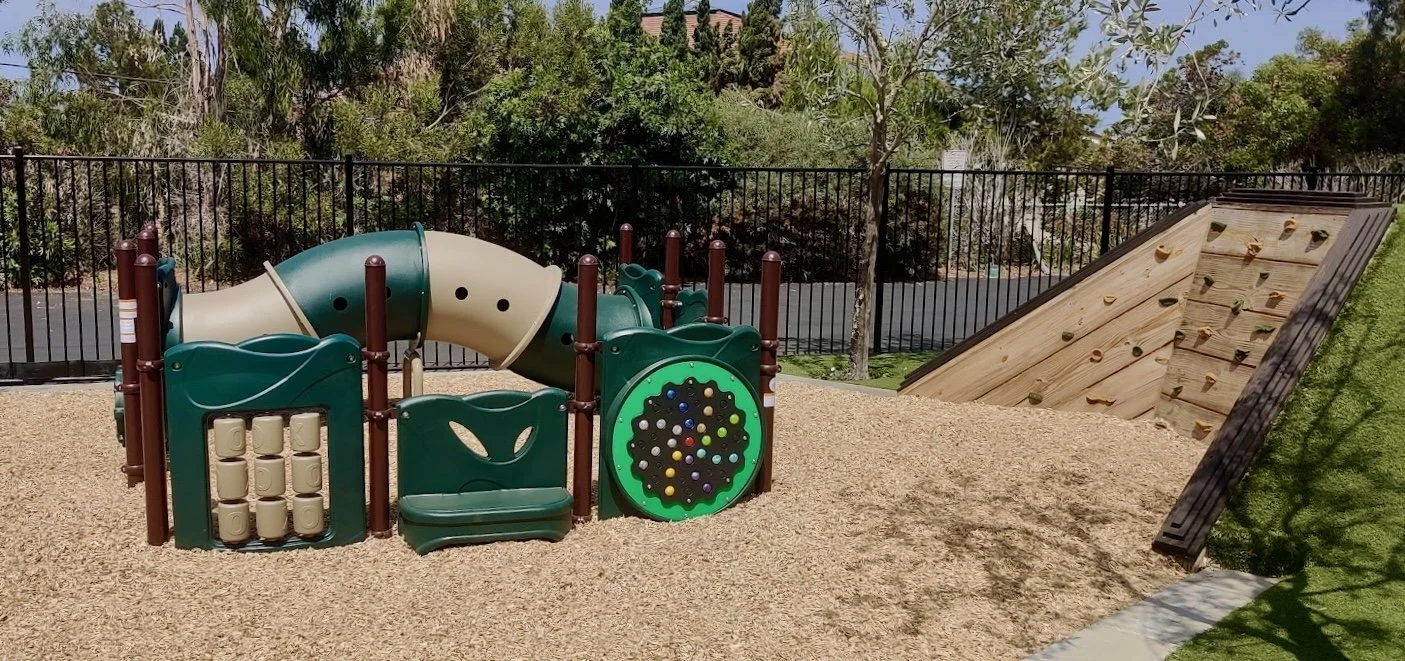Children's playground with tube slide, climbing wall, tic-tac-toe panel, and a swing set, surrounded by a black metal fence and trees.