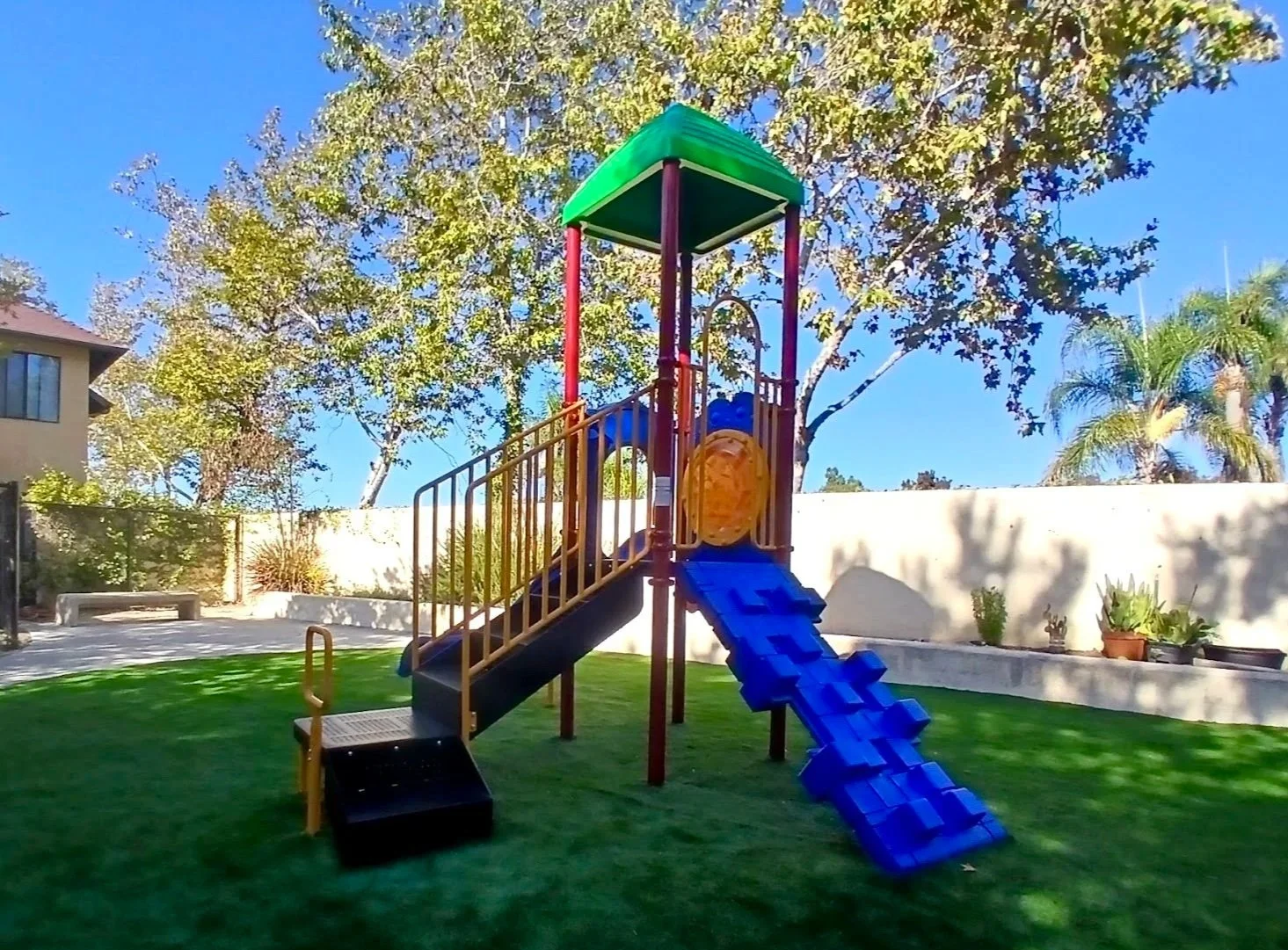 Colorful children’s playground structure with a slide, climbing ramp, and upper platform, set on a grassy yard with trees and a residential building in the background on a sunny day.