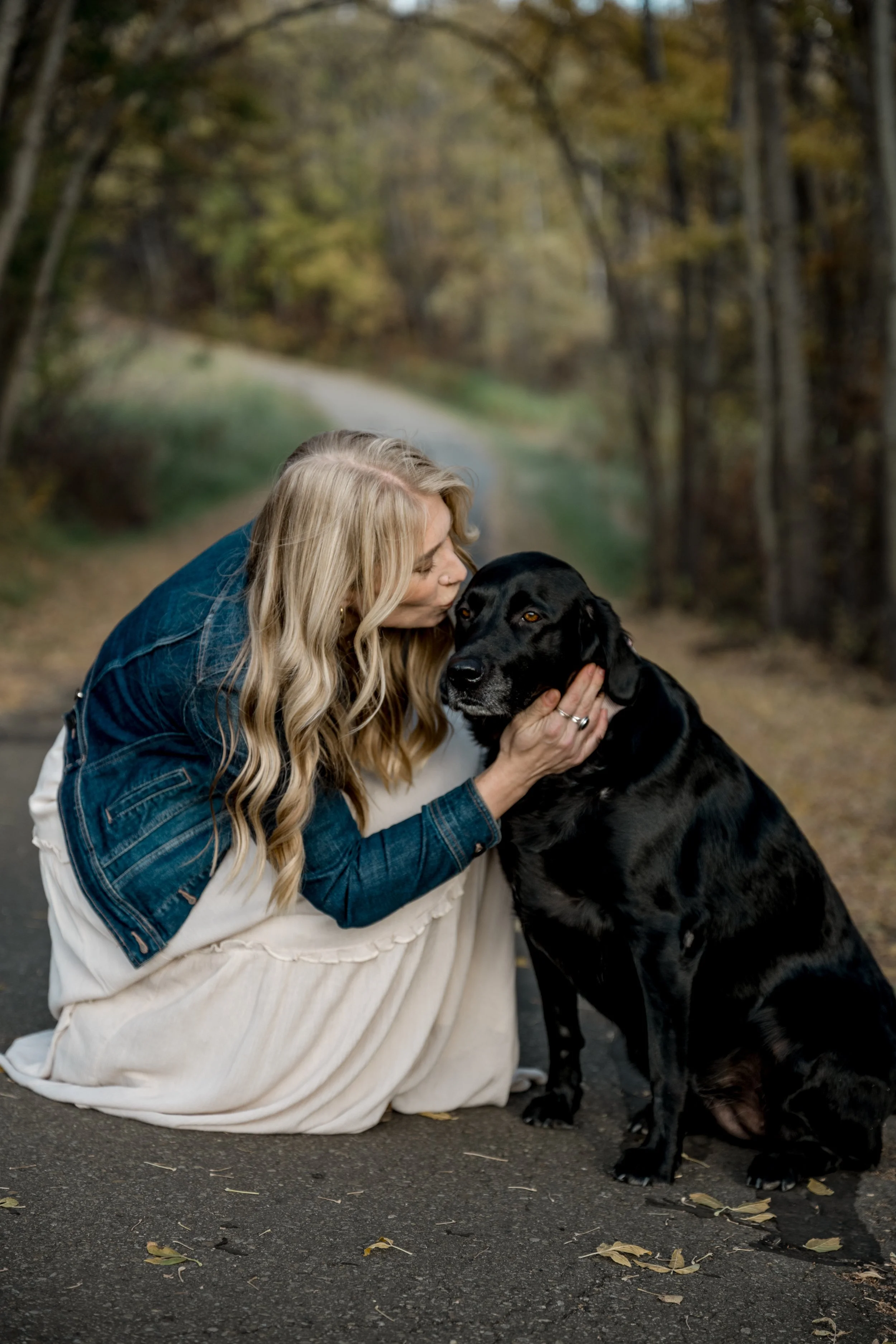 A woman with long blonde hair kneeling on a road in a forest, holding and kissing a large black dog.