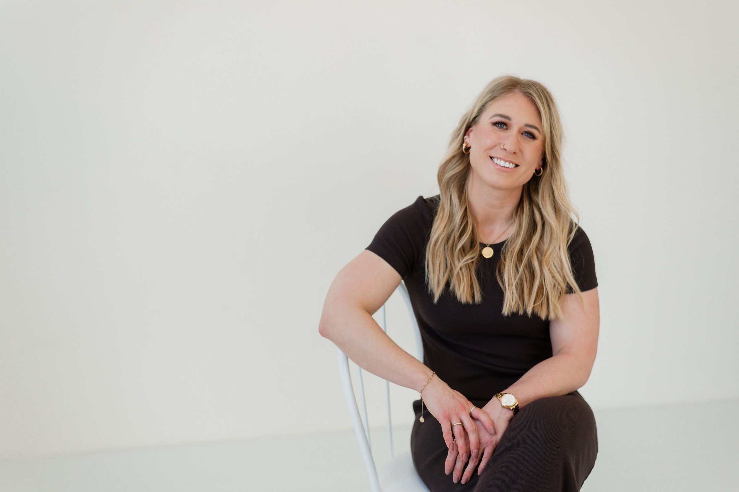 A woman with long blonde wavy hair smiling, wearing a black t-shirt, gold jewelry, and sitting on a white chair against a plain light background.
