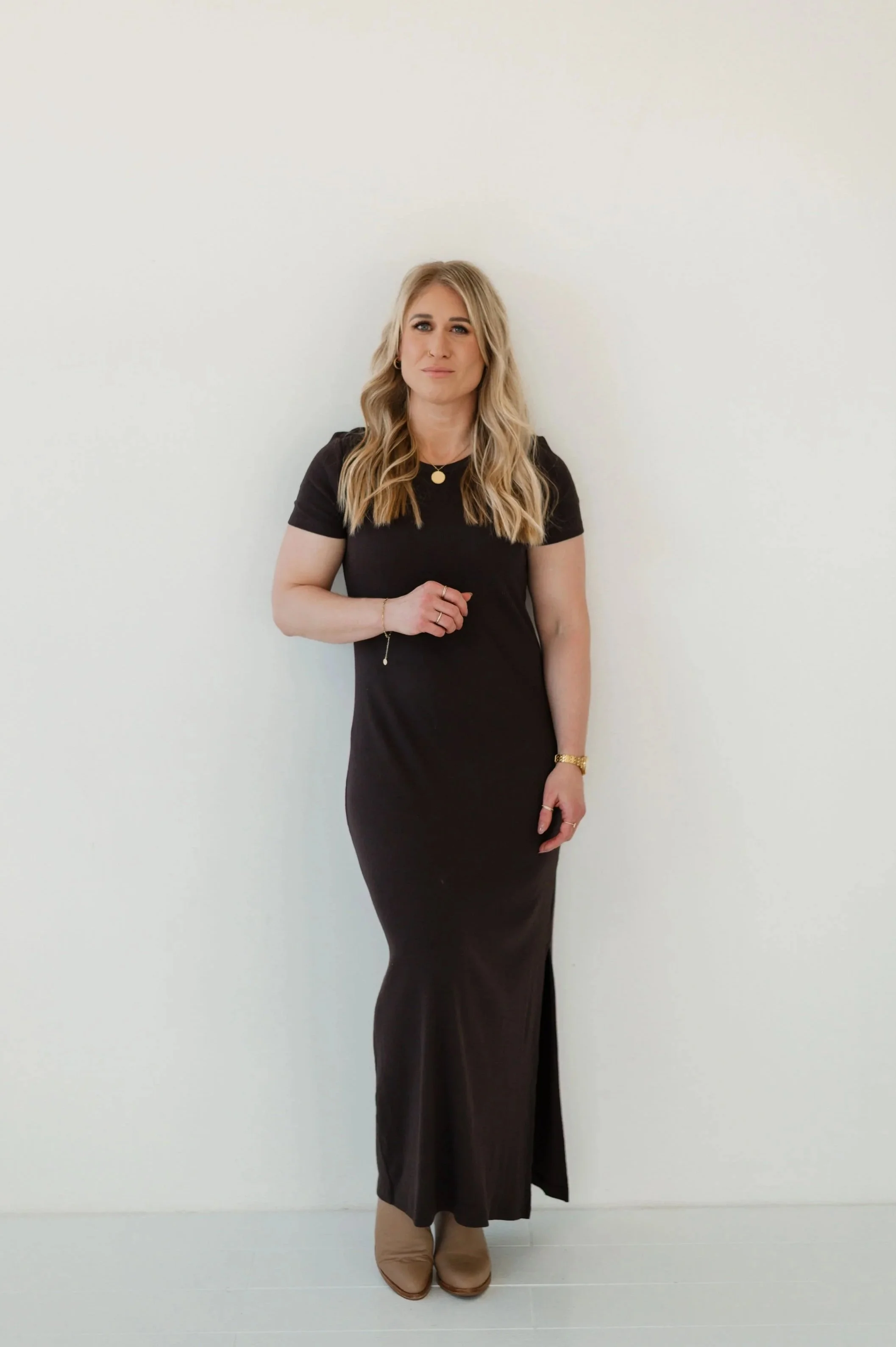 A woman with blonde hair and light skin wearing a black dress and beige ankle boots, standing against a plain white wall.