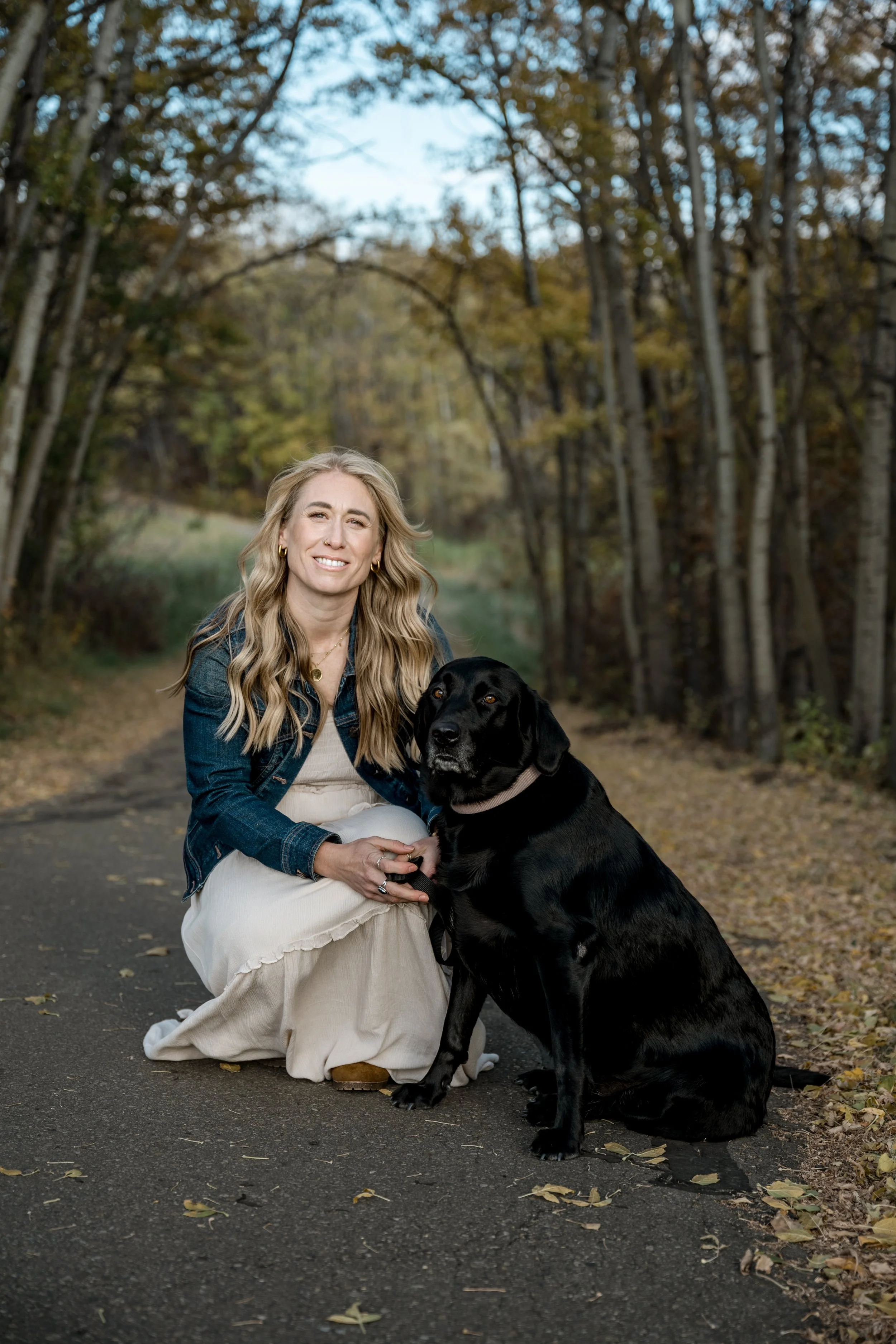 A woman with long blonde hair wearing a denim jacket and a light-colored dress kneeling on a paved path surrounded by trees with autumn leaves, holding a black Labrador retriever dog.