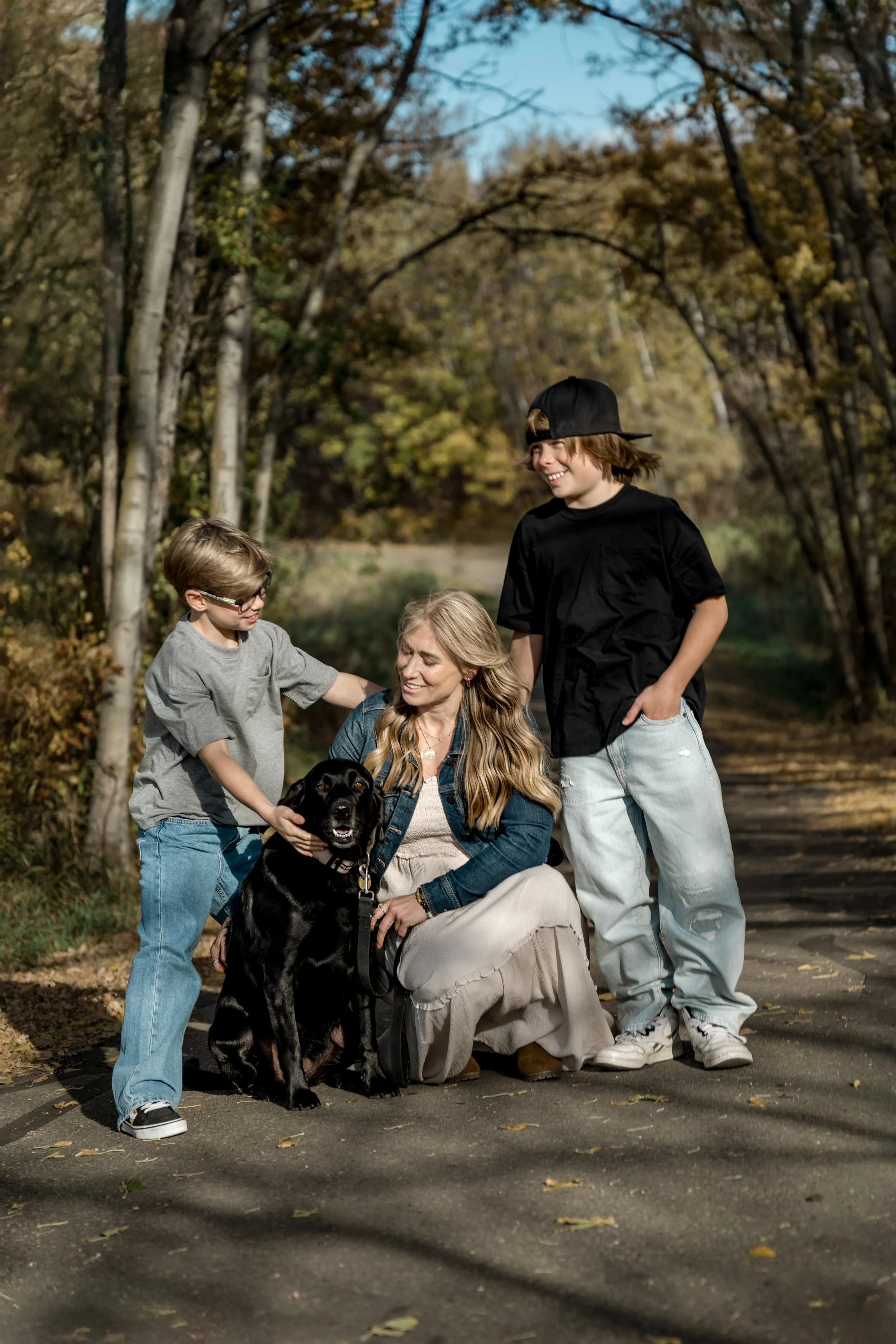 A woman and two boys smiling and playing with a black dog on a forest path during autumn.