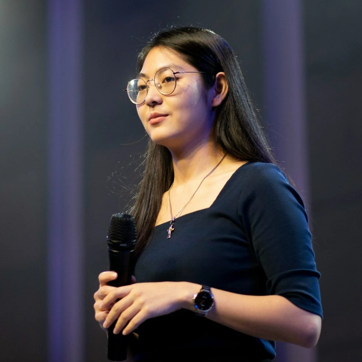 A young woman with long dark hair, glasses, and a cross necklace holds a microphone while speaking at an event.