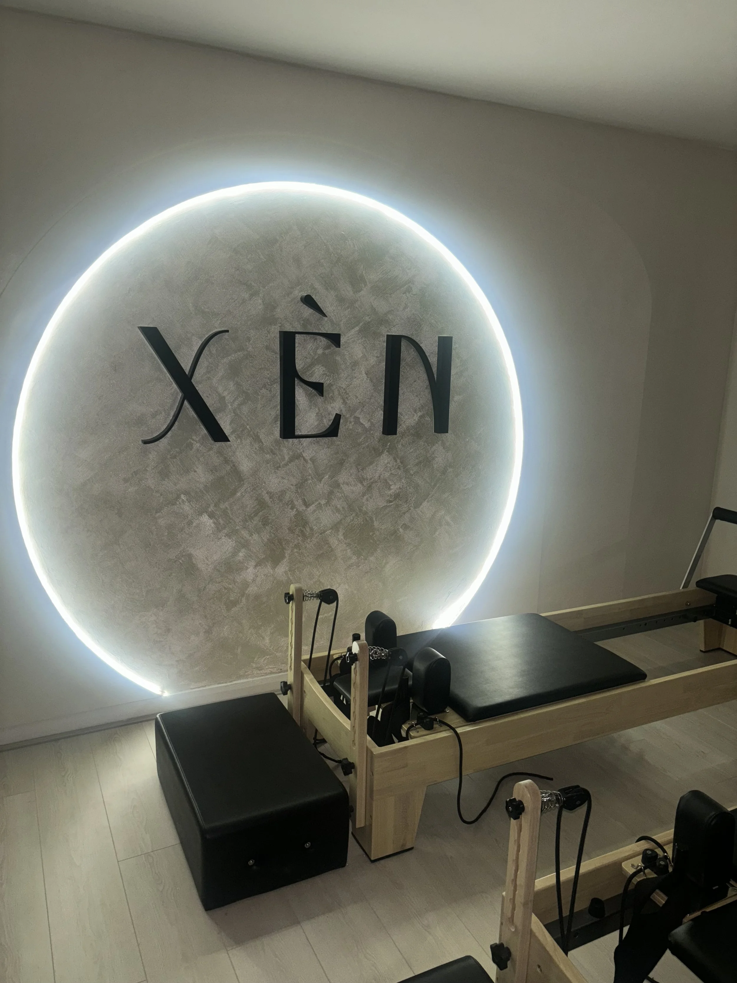 A Pilates reformer machine in front of a wall with the word 'XÉN' backlit by LED lights.