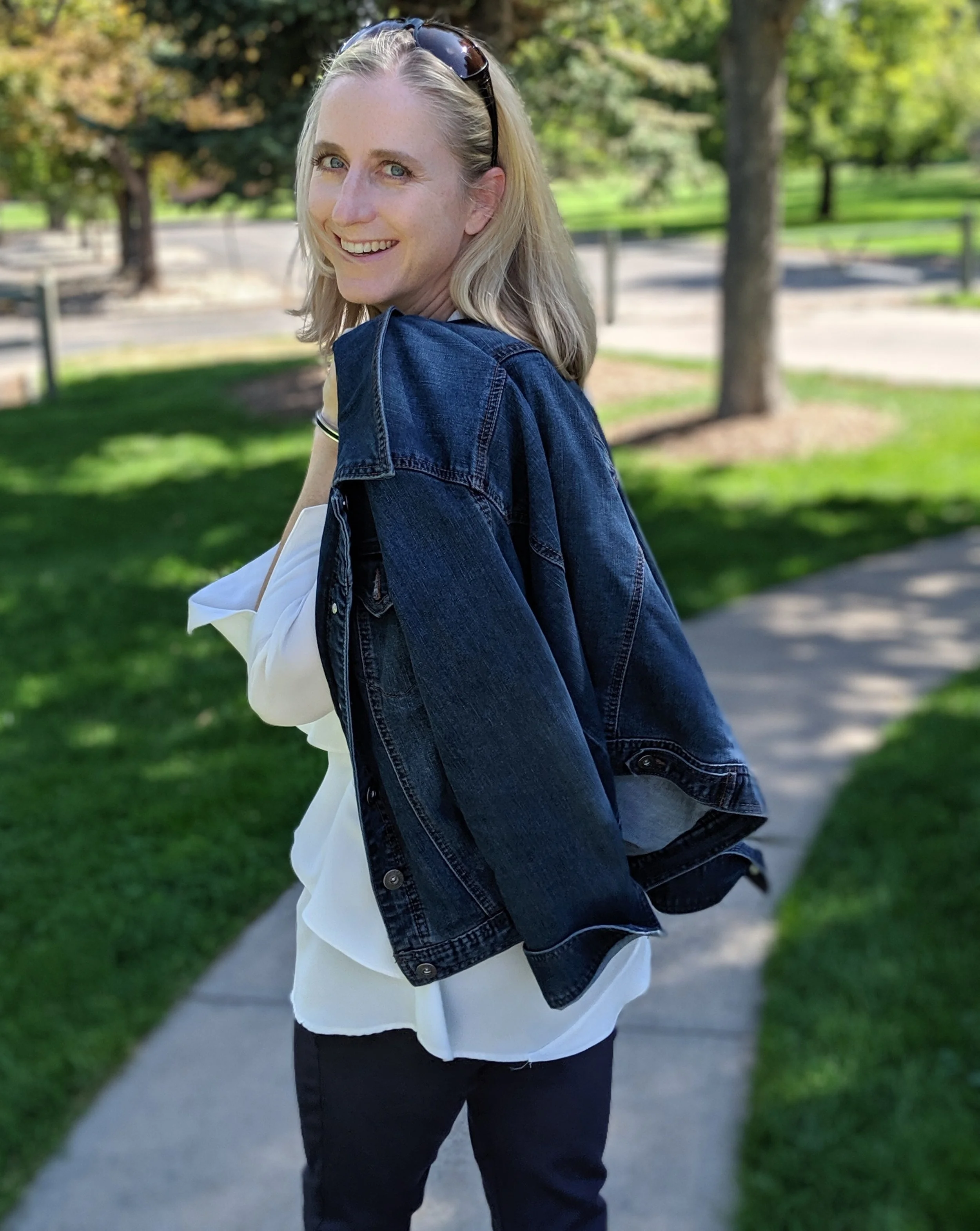 A smiling blonde woman in a white top, black pants, and a denim jacket draped over her shoulders standing on a sidewalk in a park with green grass and trees.