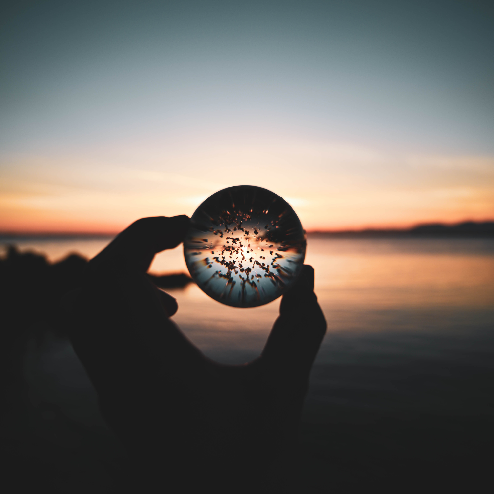 A person holding a spherical glass object with a sunset or sunrise in the background, overlooking a body of water.