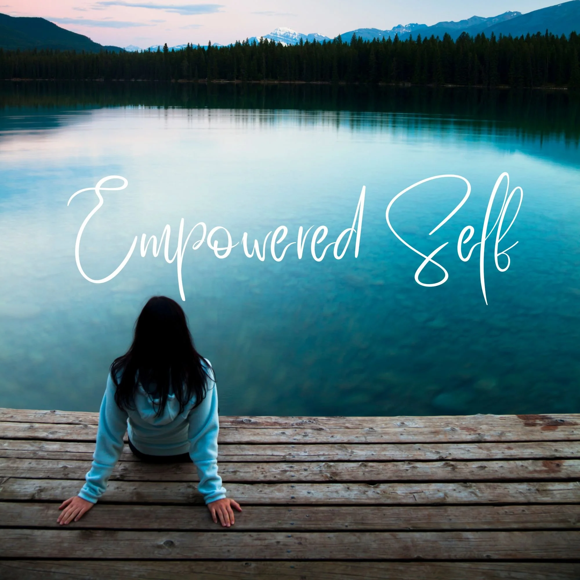 A person in a white hoodie and light blue pants crouching on a wooden dock, facing a large mountain lake with pine trees and snow-capped mountains in the background, during early evening with a cloudy sky.
