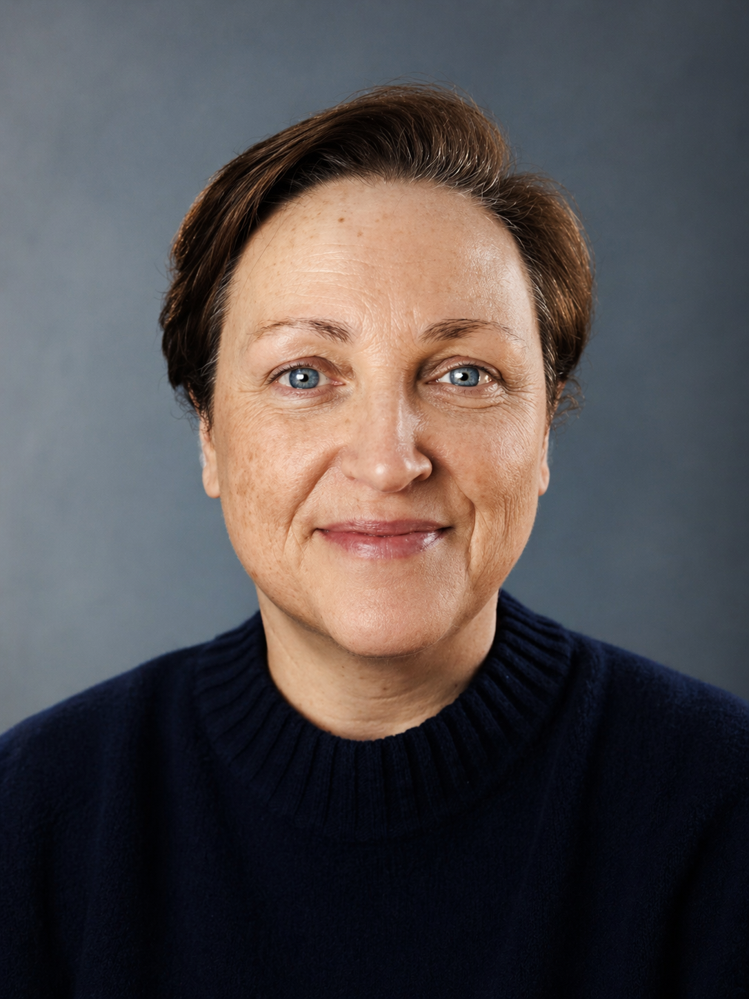 A middle-aged woman with short brown hair and blue eyes smiling, wearing a dark navy sweater against a plain gray background.