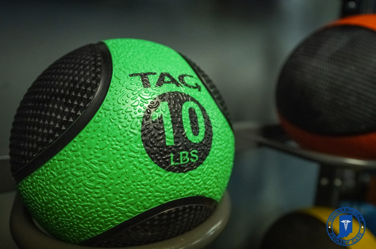 A close-up of a green and black medicine ball labeled 'TAG 10 LBS' with textured surface, placed on a rack in a gym or medical facility. Other medicine balls are partially visible in the background.