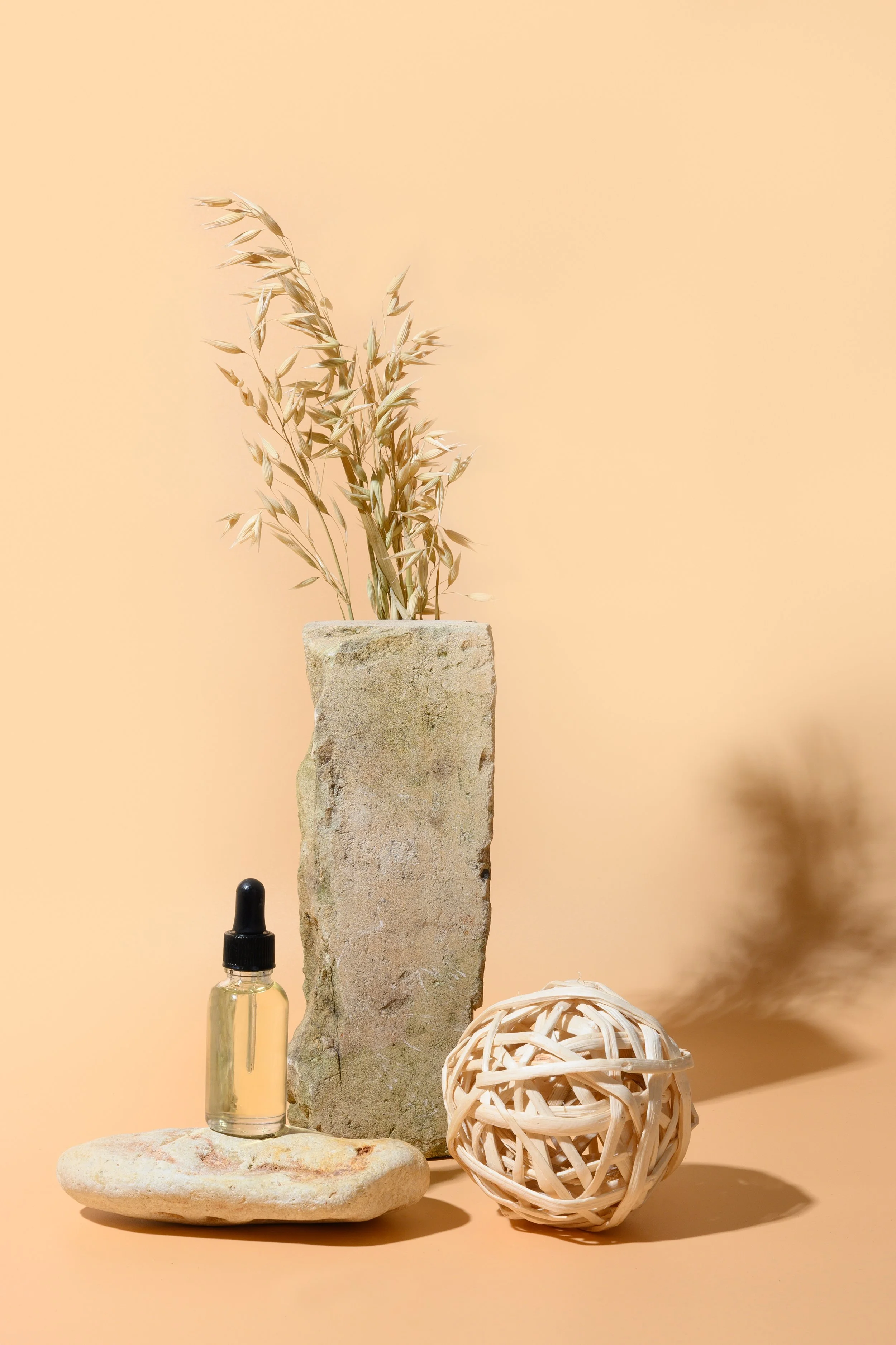 Arrangement of decorative objects including a small glass dropper bottle, a large stone, a tall rough stone with dried beige grass, and a woven rattan ball on a beige surface against a beige background.