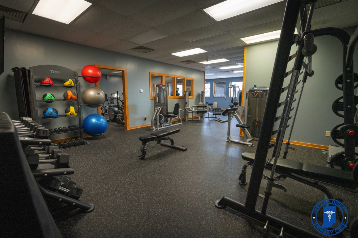 An empty gym with various workout equipment including dumbbells, medicine balls, exercise benches, and cardio machines. The gym has blue walls, windows, and a black rubberized floor.