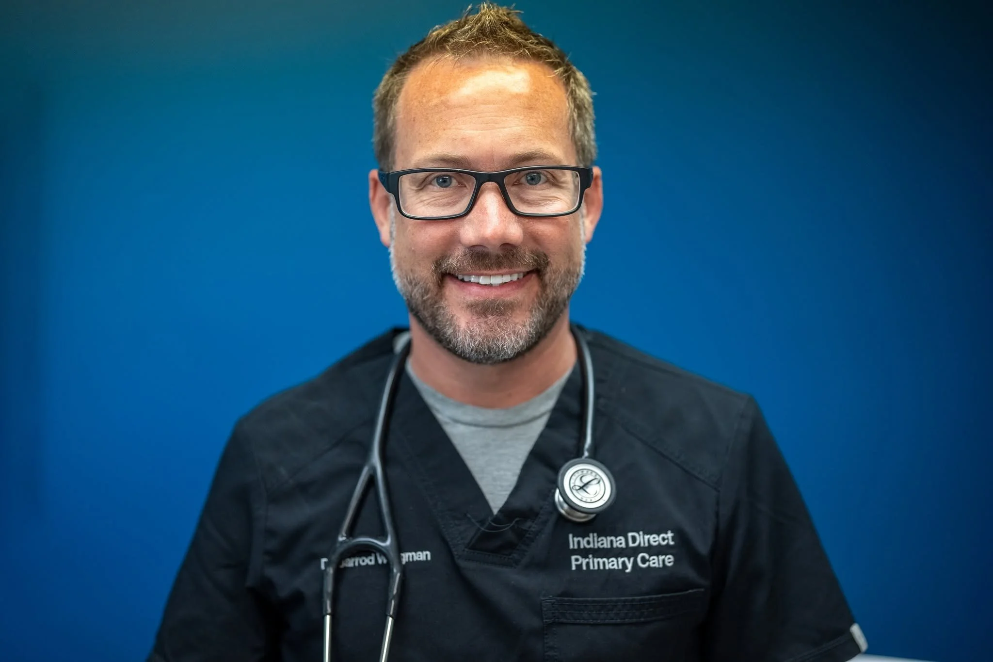 Portrait of a male medical professional wearing glasses and a stethoscope around his neck, smiling at the camera, against a blue background.