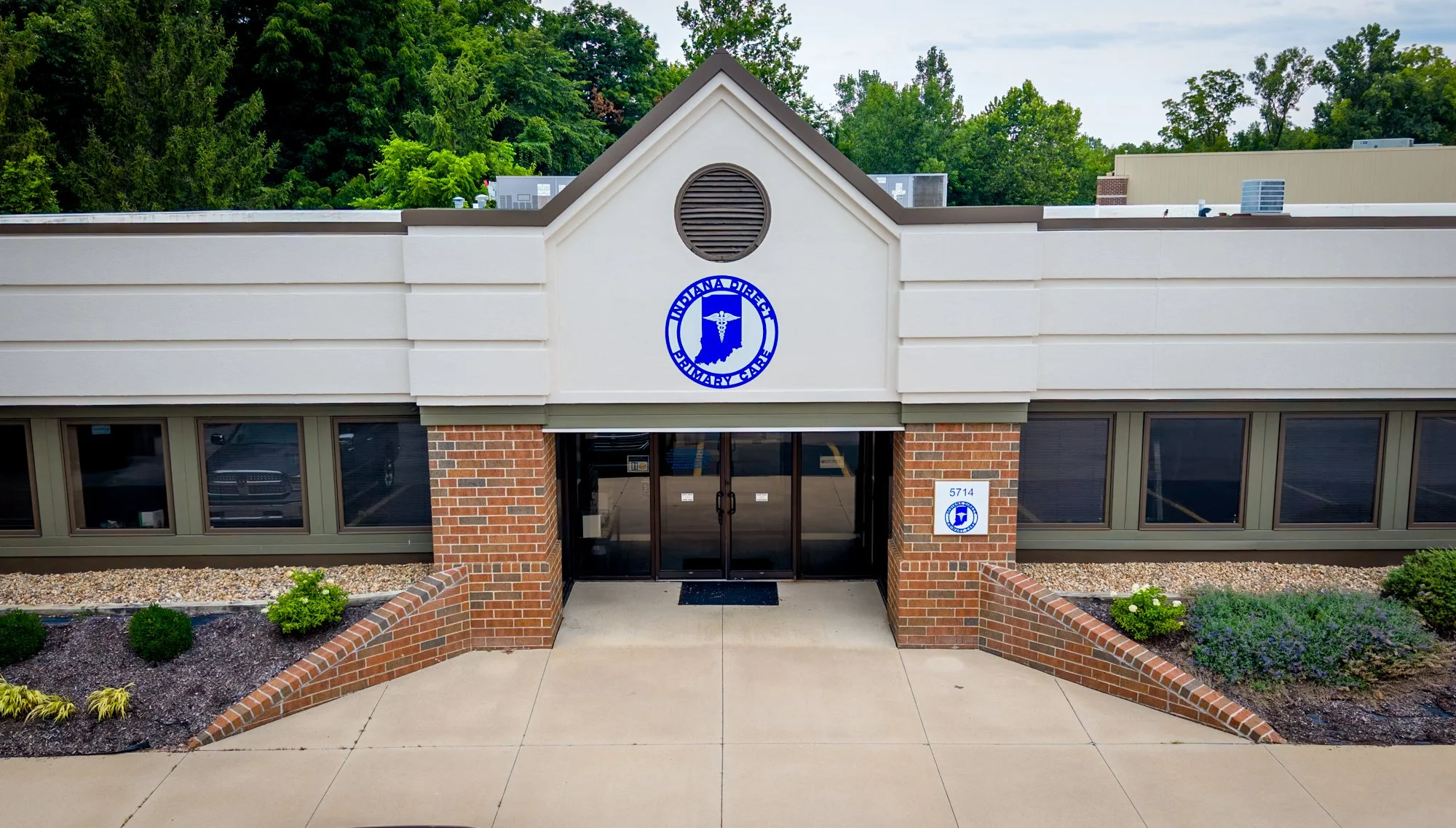 Front entrance of a building with a sign that reads 'Police Public' and an emblem featuring a medical symbol. The building has brick pillars, large windows, and a landscaped area with small plants and flowers.