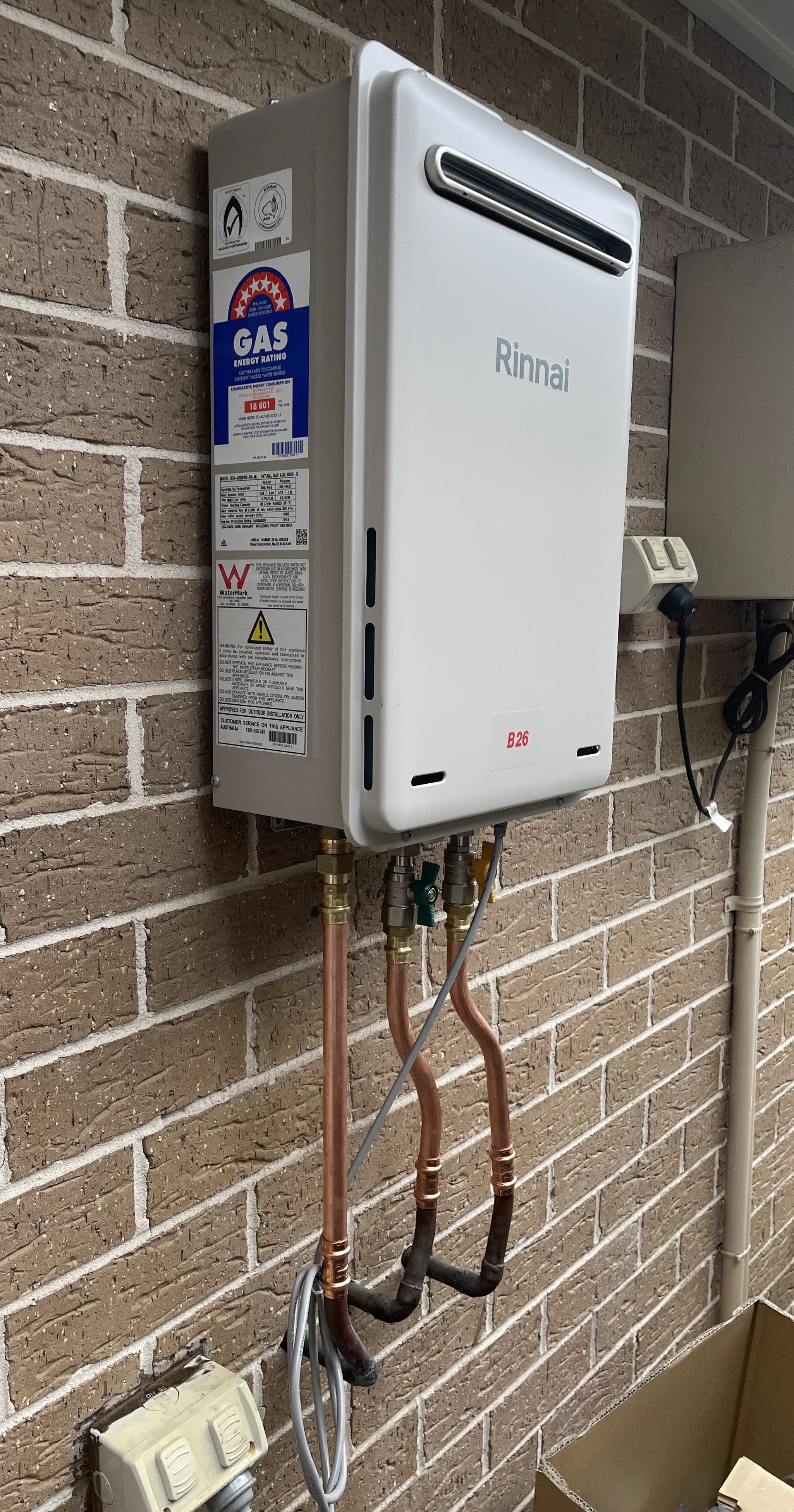 An outdoor Rinnai tankless water heater mounted on a brick wall with copper pipes, electrical connections, and labels.