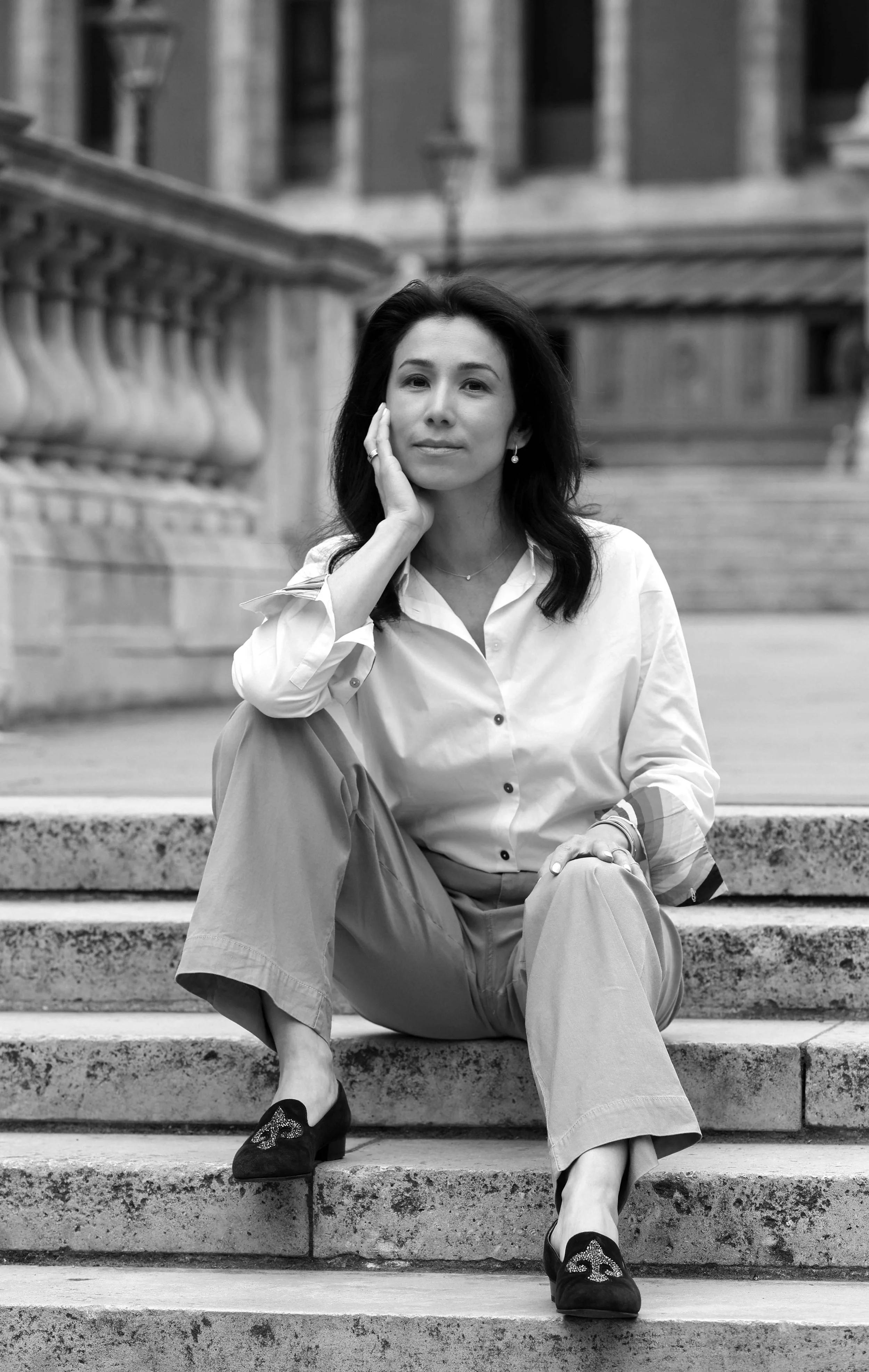 Black and white photo of a woman sitting on outdoor stone steps, wearing a button-up shirt, wide-leg pants, and heels, with a hand resting on her face, in front of a decorative stone railing and a building in the background.