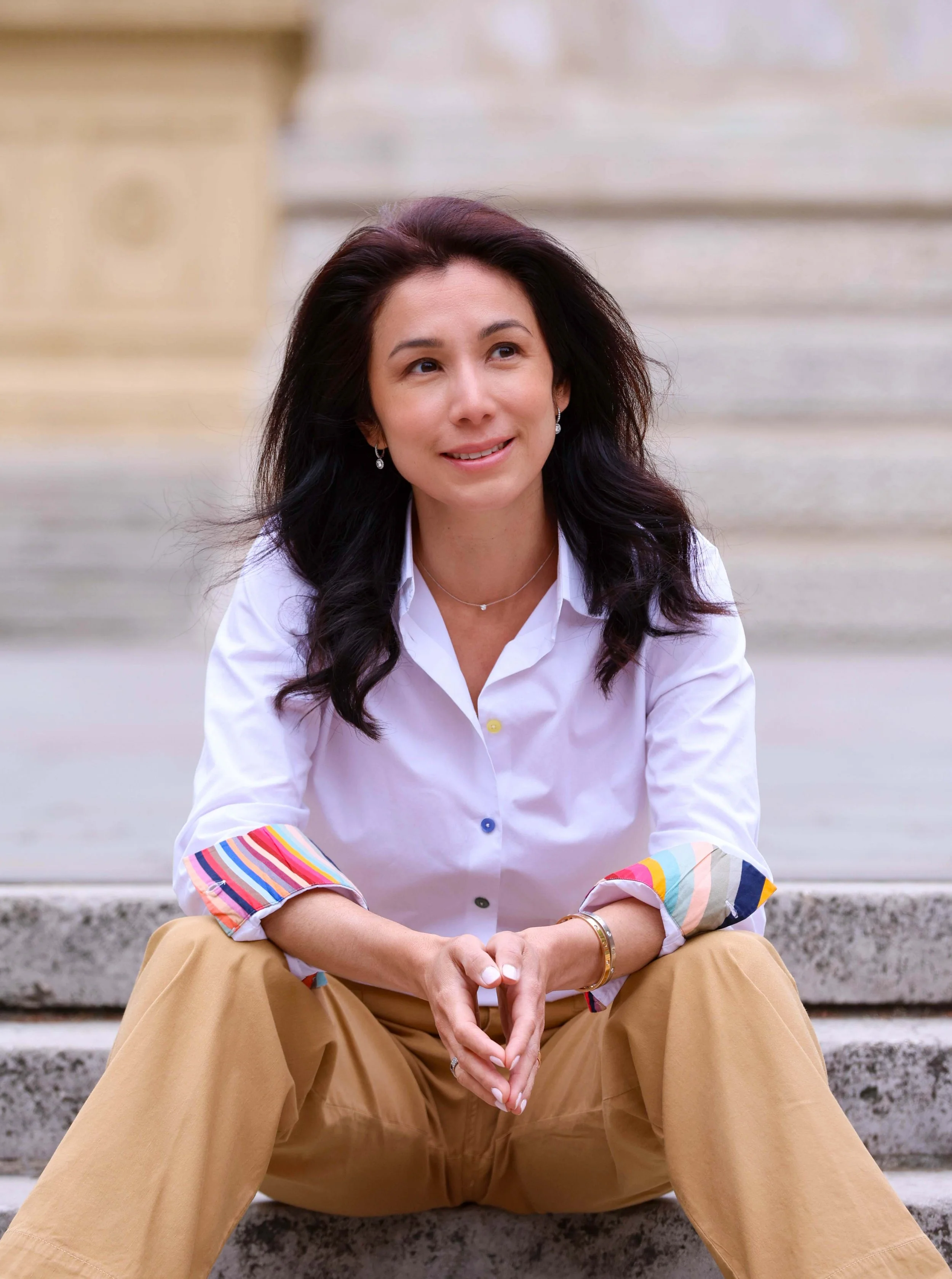 A woman sitting on concrete steps outdoors, smiling and looking to the side. She has dark, wavy hair and is wearing a white shirt with colorful striped cuffs, beige pants, earrings, a necklace, and a bracelet.
