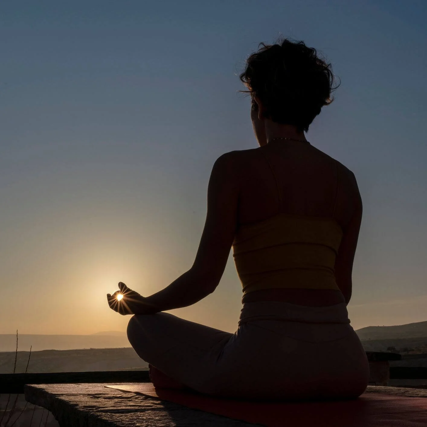 A person practicing yoga or meditation outdoors at sunset, sitting cross-legged on a mat with their hand holding the sun.