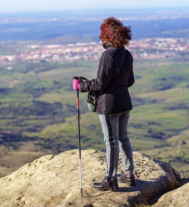 A woman standing on a rocky hilltop with a hiking pole, overlooking a green landscape and a distant town under a blue sky.