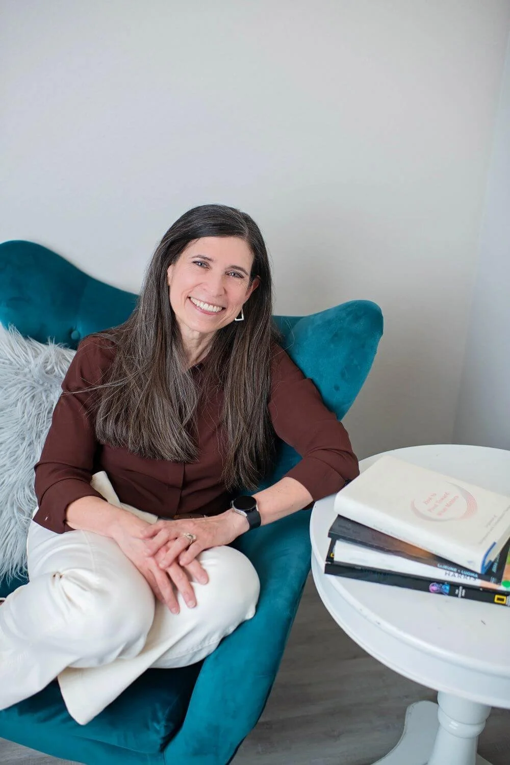 A woman with long dark hair, wearing a brown top and white pants, sitting on a teal sofa with a fluffy white pillow. She is smiling and looking at the camera. There is a round white table beside her with some books and papers on it.