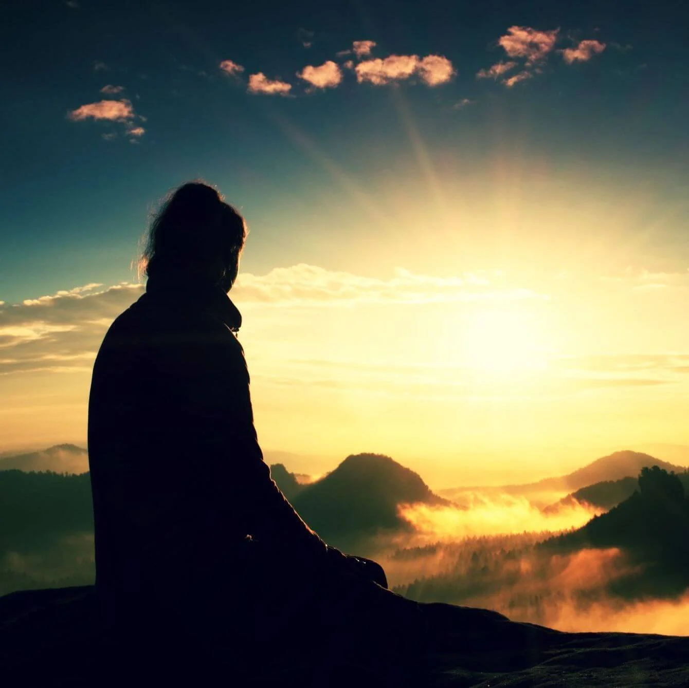 A silhouette of a person sitting on a rock watching the sunrise over mountains and clouds.