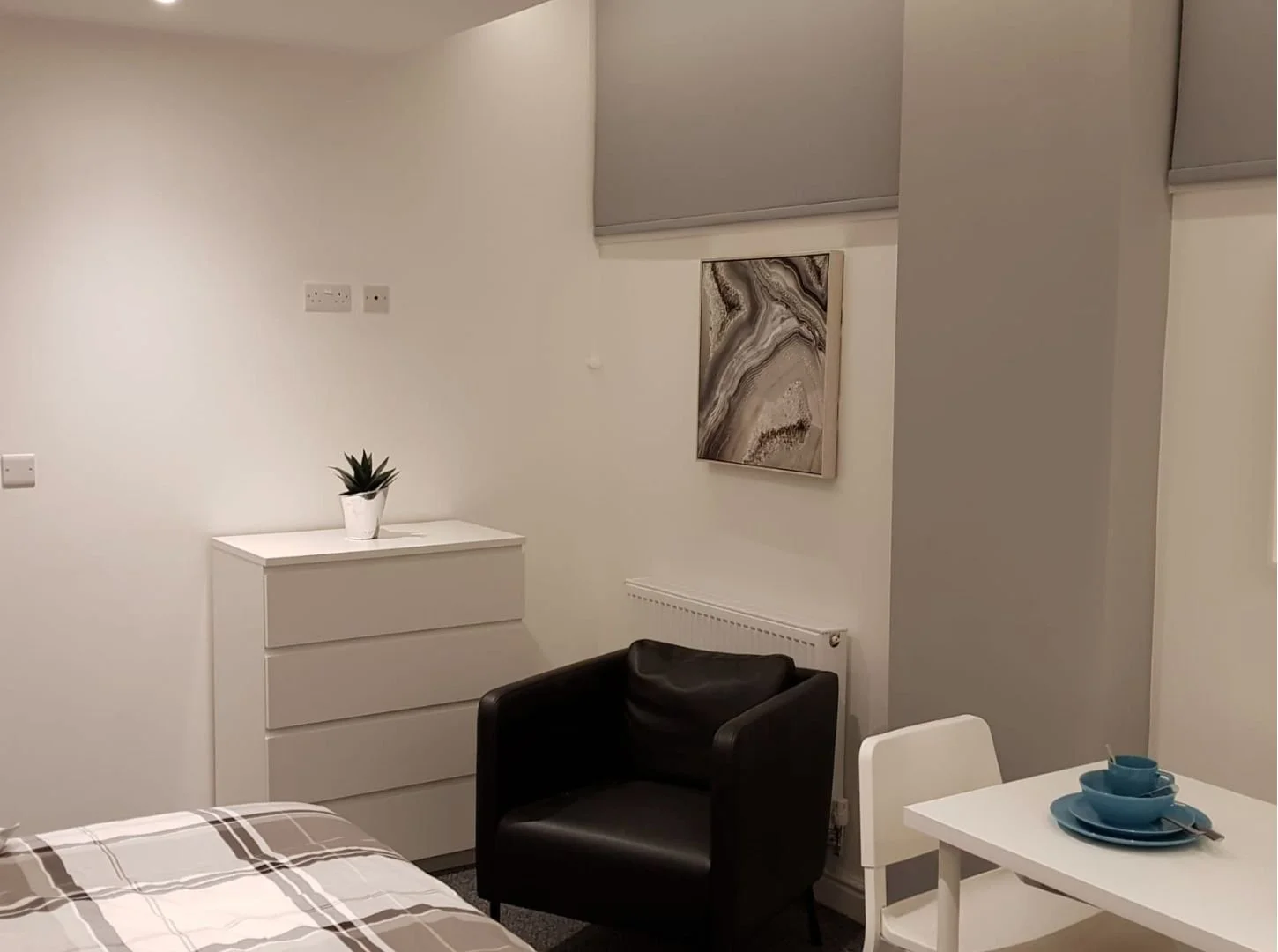A minimalist bedroom corner with a white dresser, a potted plant on top, a black armchair, a small white dining table with blue bowls and cups, a white chair, and a framed abstract art piece on the wall.