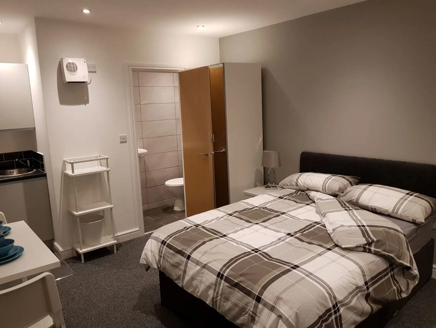 A cozy bedroom with a bed featuring plaid bedding, a bedside table with a lamp, a wardrobe, and a small kitchenette area. There is an open door leading to a bathroom with tiled walls and a toilet.