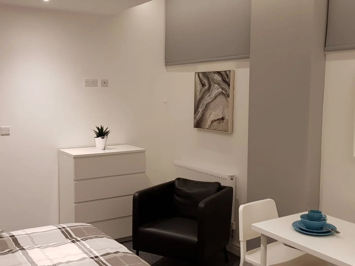 Interior of a small room with white walls, a white dresser with a potted succulent, a black armchair, a white table with blue dishes, and wall art.