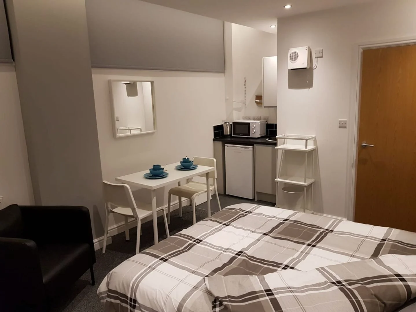 A small studio apartment with a bed, a black armchair, a white dining table with two chairs set with blue dishes, a compact kitchen with appliances, and a white shelving unit against a white wall.