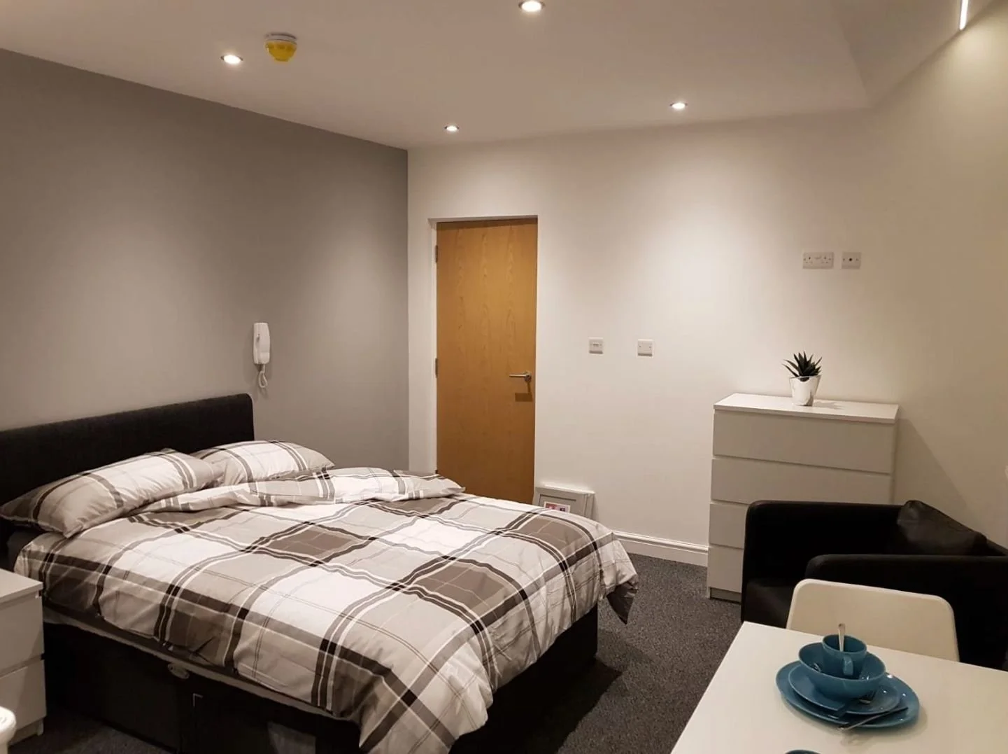 Modern bedroom with a bed, black armchair, white dresser with a plant, and white table with blue dishes