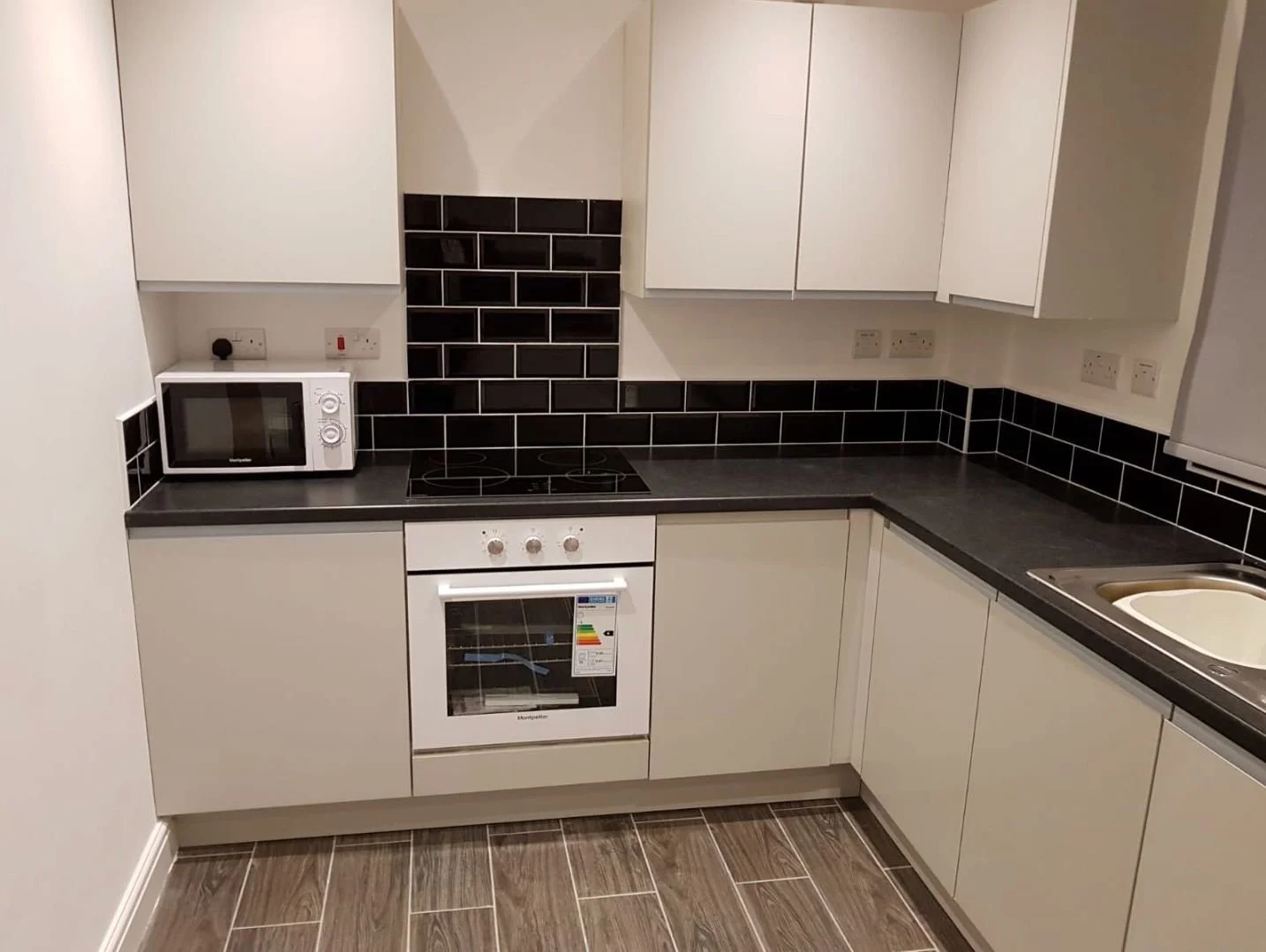 A compact kitchen with white cabinets, black countertop, black tiled backsplash, a white microwave, an electric stove, an oven, and a sink. The floor has wood-look tile.