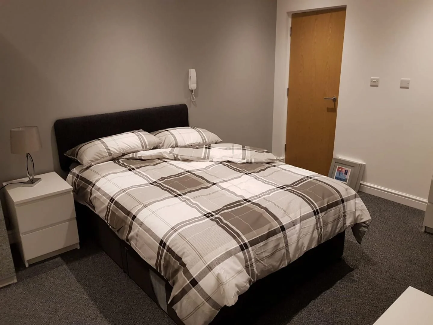 A bedroom with a bed featuring a black headboard, covered with beige, white, and brown plaid bedding and matching pillows. To the left of the bed is a white nightstand with a beige lamp, and on the right is a wooden door. A framed picture is leaning