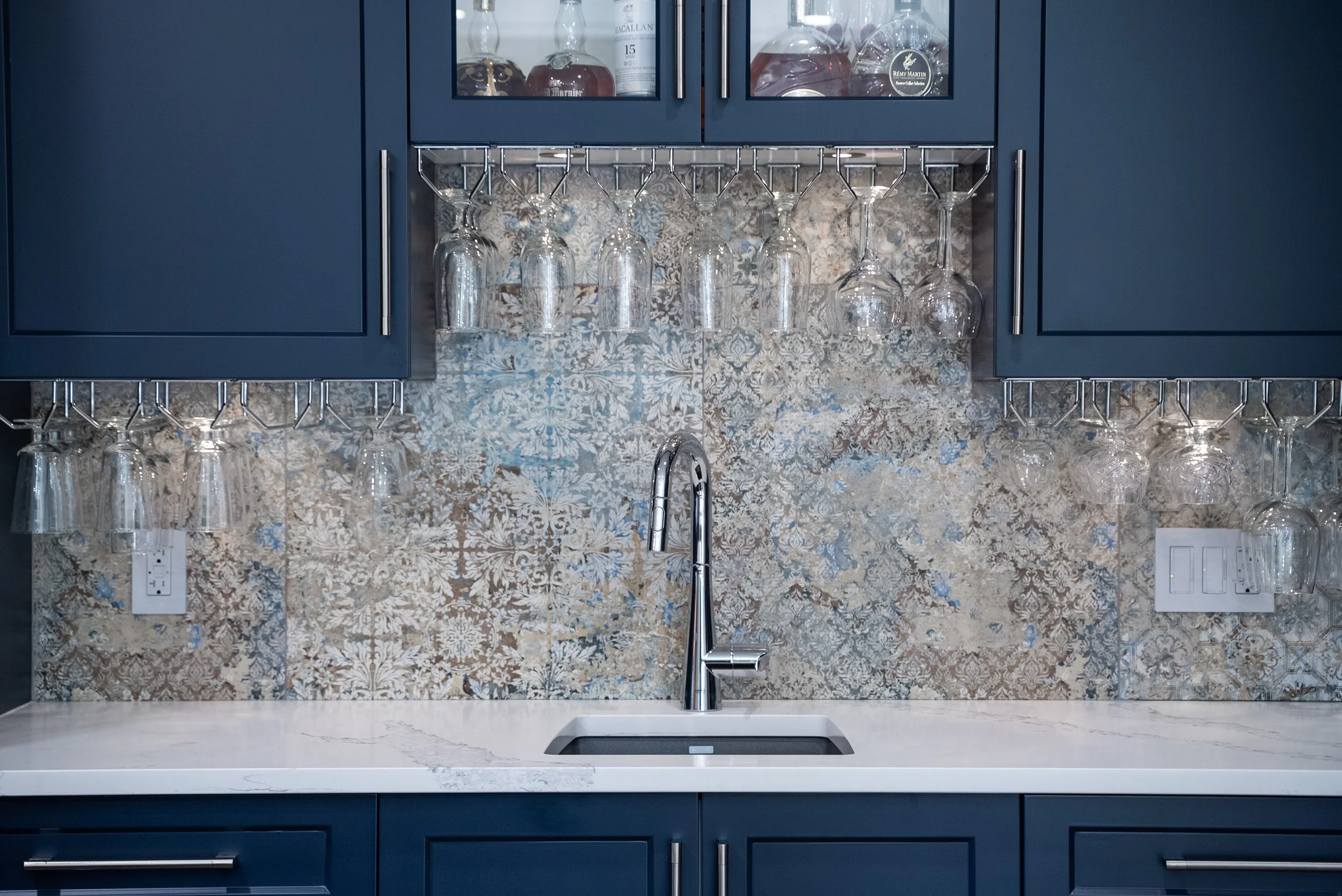 Modern kitchen with blue cabinets and a white marble countertop, glass cabinets with liquor bottles, hanging wine glasses, decorative tiled backsplash, and a sleek chrome faucet.