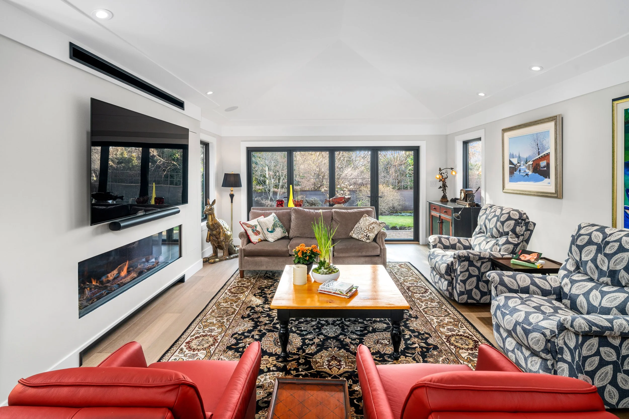 Living room with a fireplace, flat-screen TV, large sliding glass doors leading to a backyard, and various furniture including armchairs, a sofa, a coffee table with plants and books, and decorative artwork.