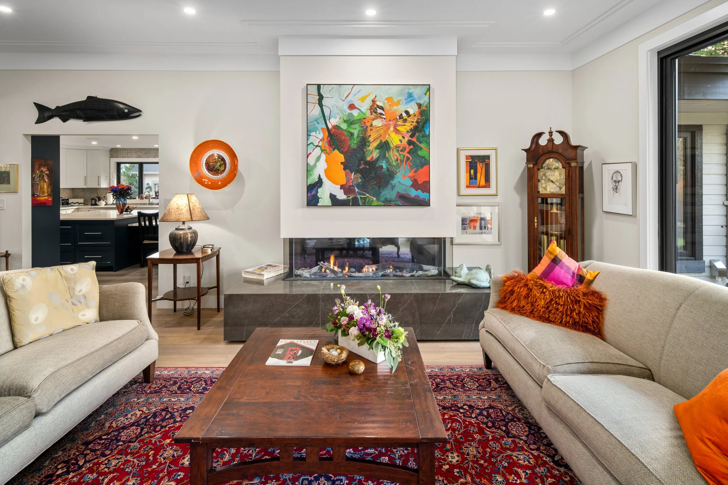 Living room with beige sofas, a wooden coffee table with a flower arrangement, colorful pillows, a fireplace with an abstract painting above it, and various artworks on the walls. A window reveals outdoor scenery.