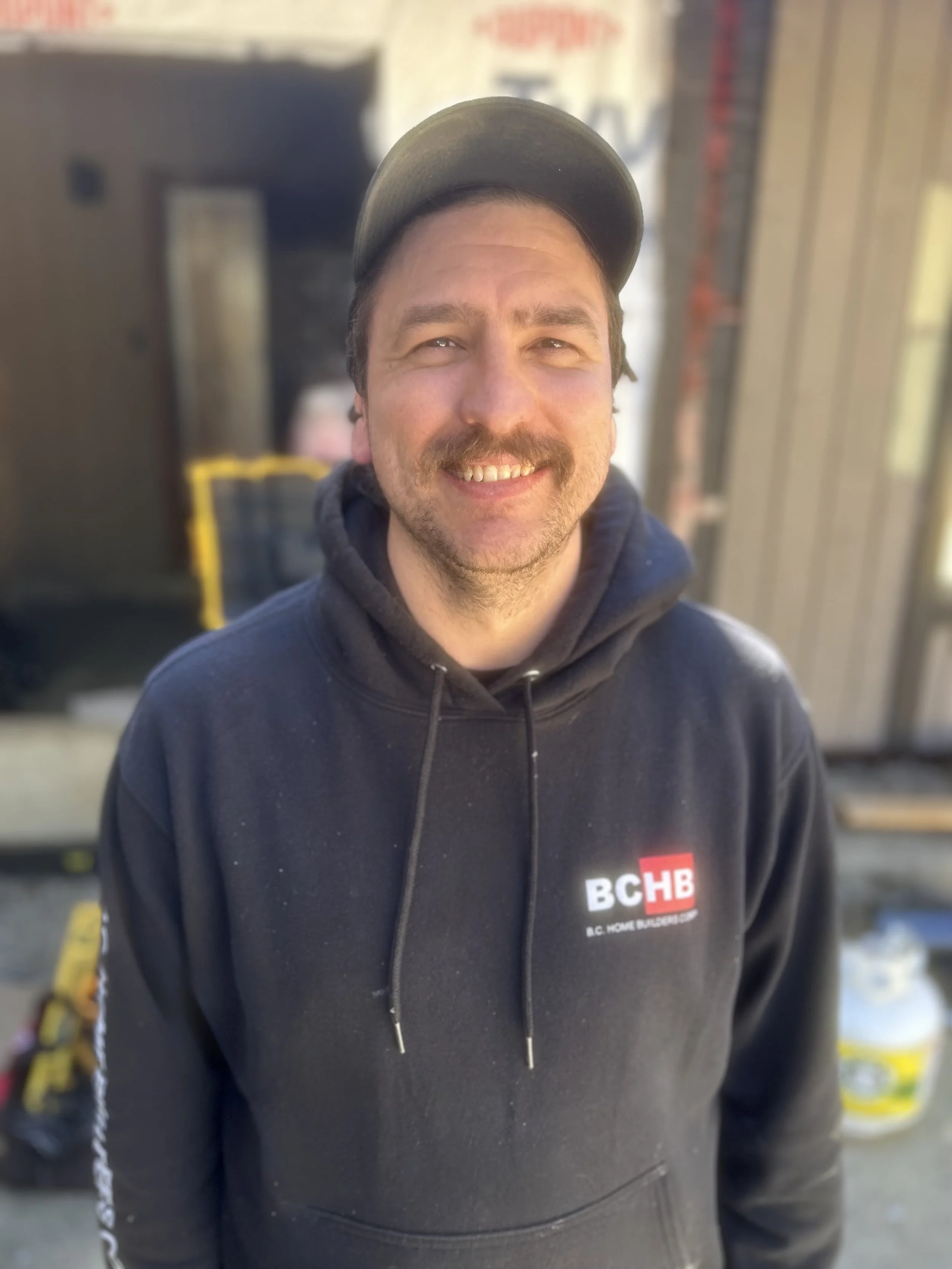 A man smiling wearing a black BC Home Builders hoodie and a cap, standing outdoors with a blurred background that appears to be a construction or work site.