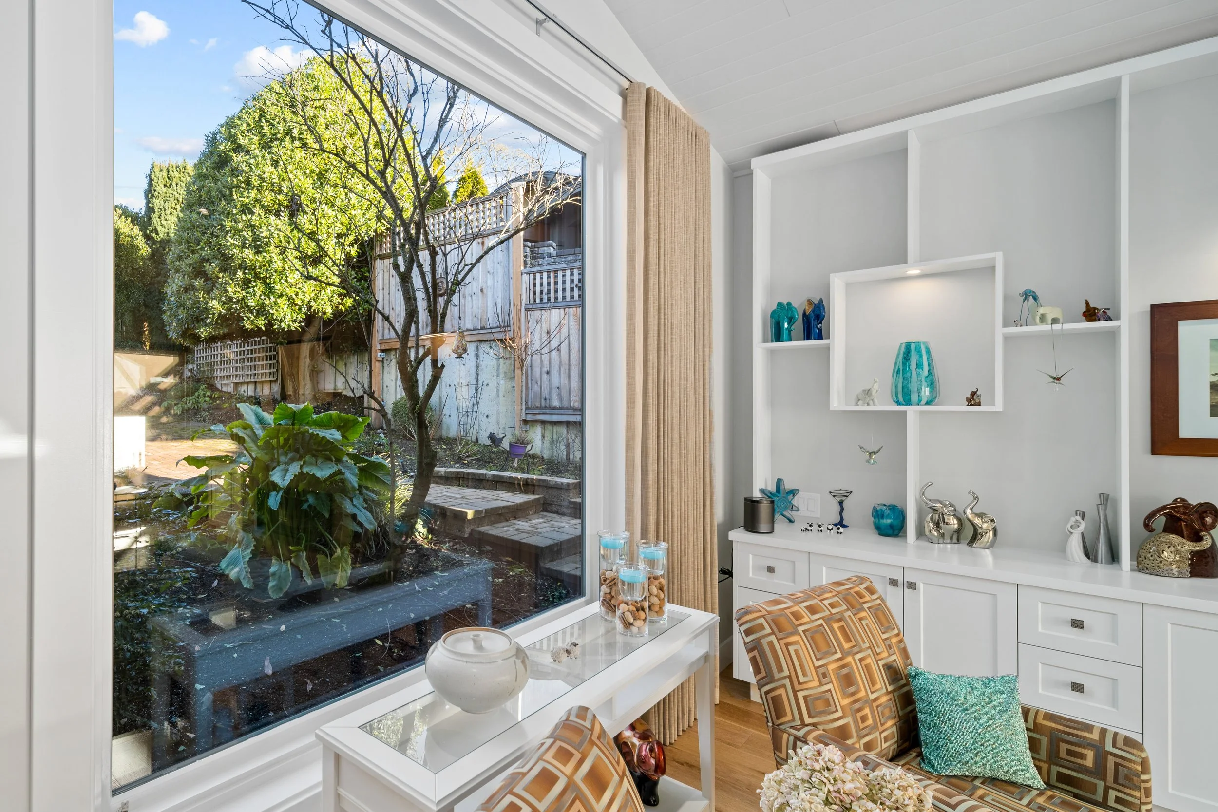 Living room with a large window showing a backyard with trees and a wooden fence, white interior walls, built-in shelves decorated with blue and silver ornaments, a patterned armchair with a teal cushion, and a side table with decorative items.