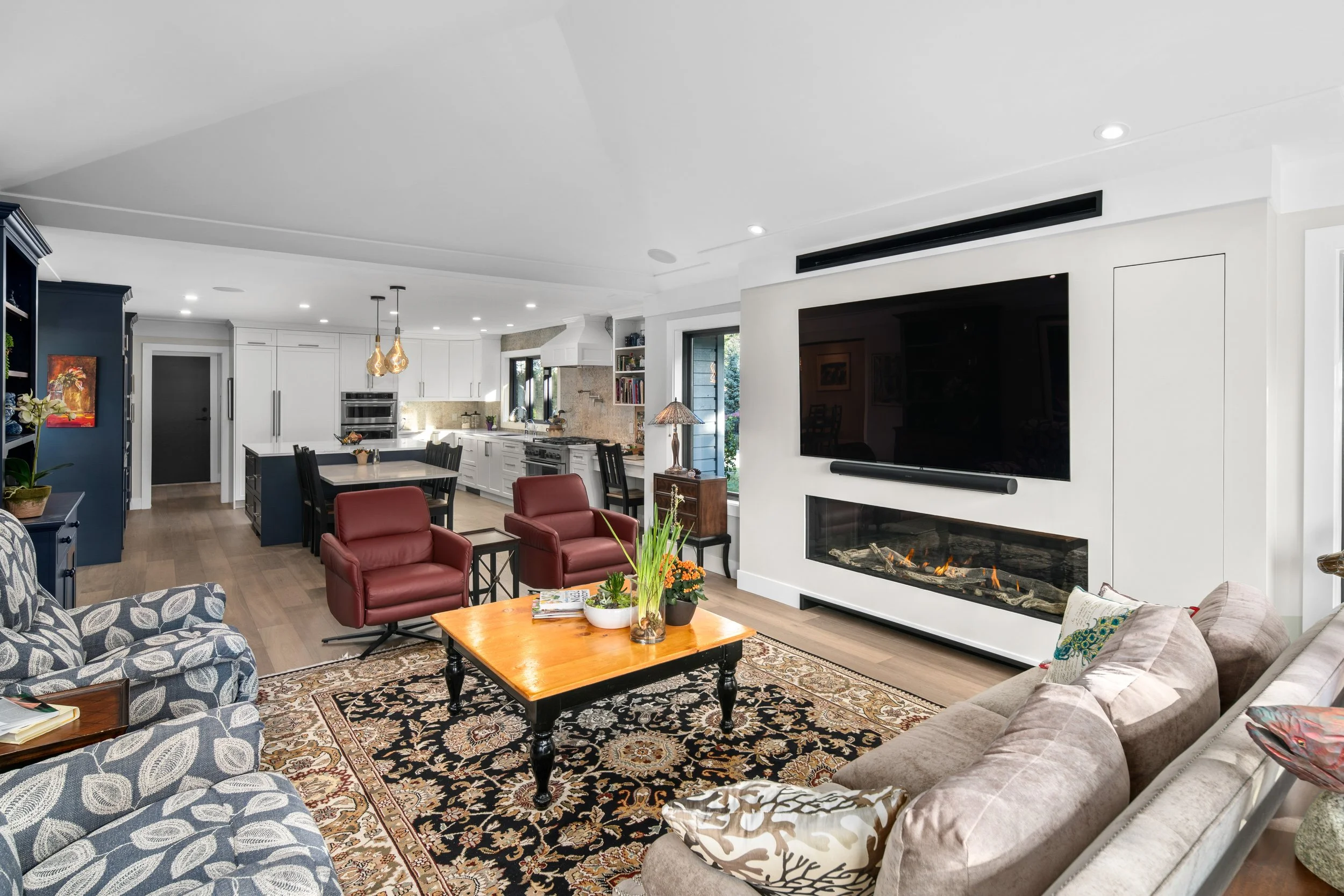 A spacious open-concept living room and kitchen with a modern design. The living room features a beige sofa with patterned pillows, two red armchairs, a wooden coffee table with plants, a patterned area rug, and a wall-mounted TV over a fireplace. The kitchen includes white cabinetry, a large island with black stools, pendant lighting, and stainless steel appliances, with large windows and hardwood flooring throughout.