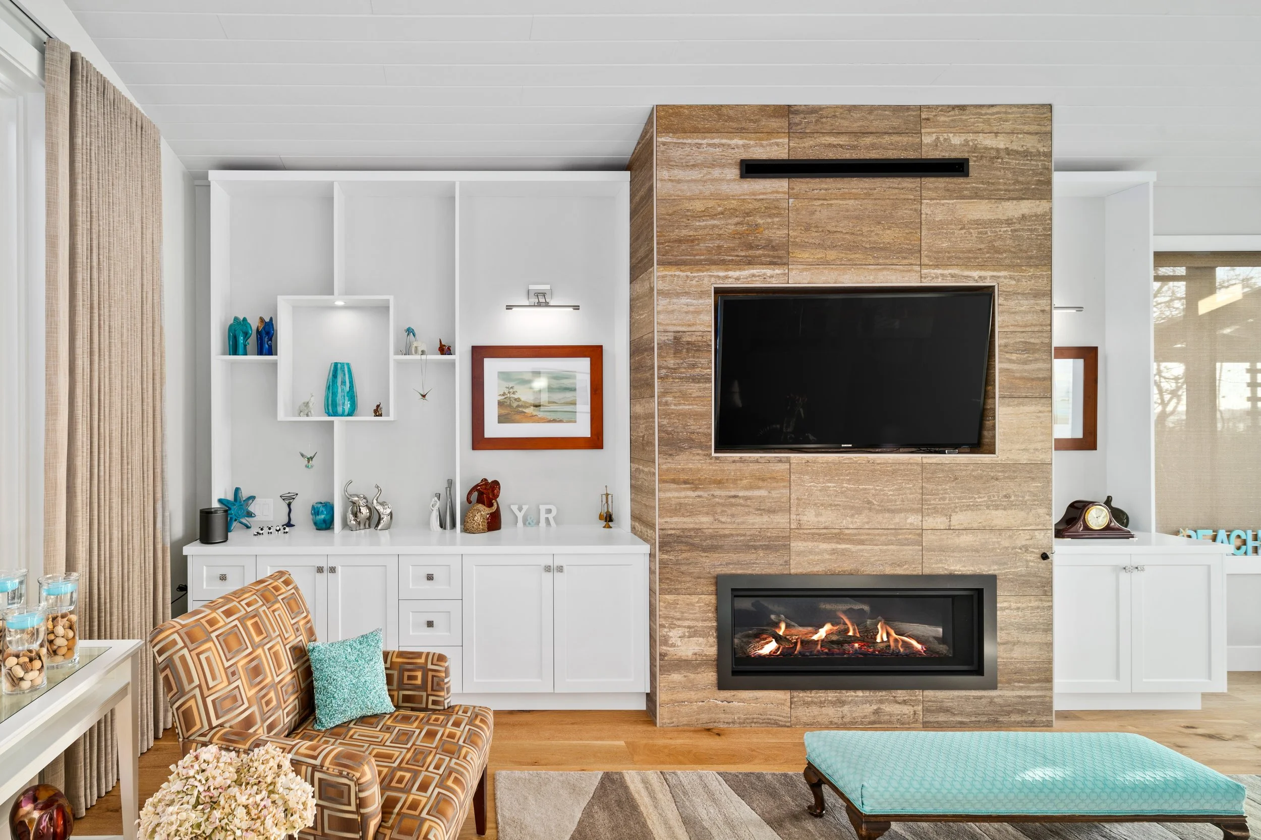 Living room with a fireplace, mounted TV, and white shelving with decorative items, an armchair with a patterned cushion, and a bench with teal upholstery.