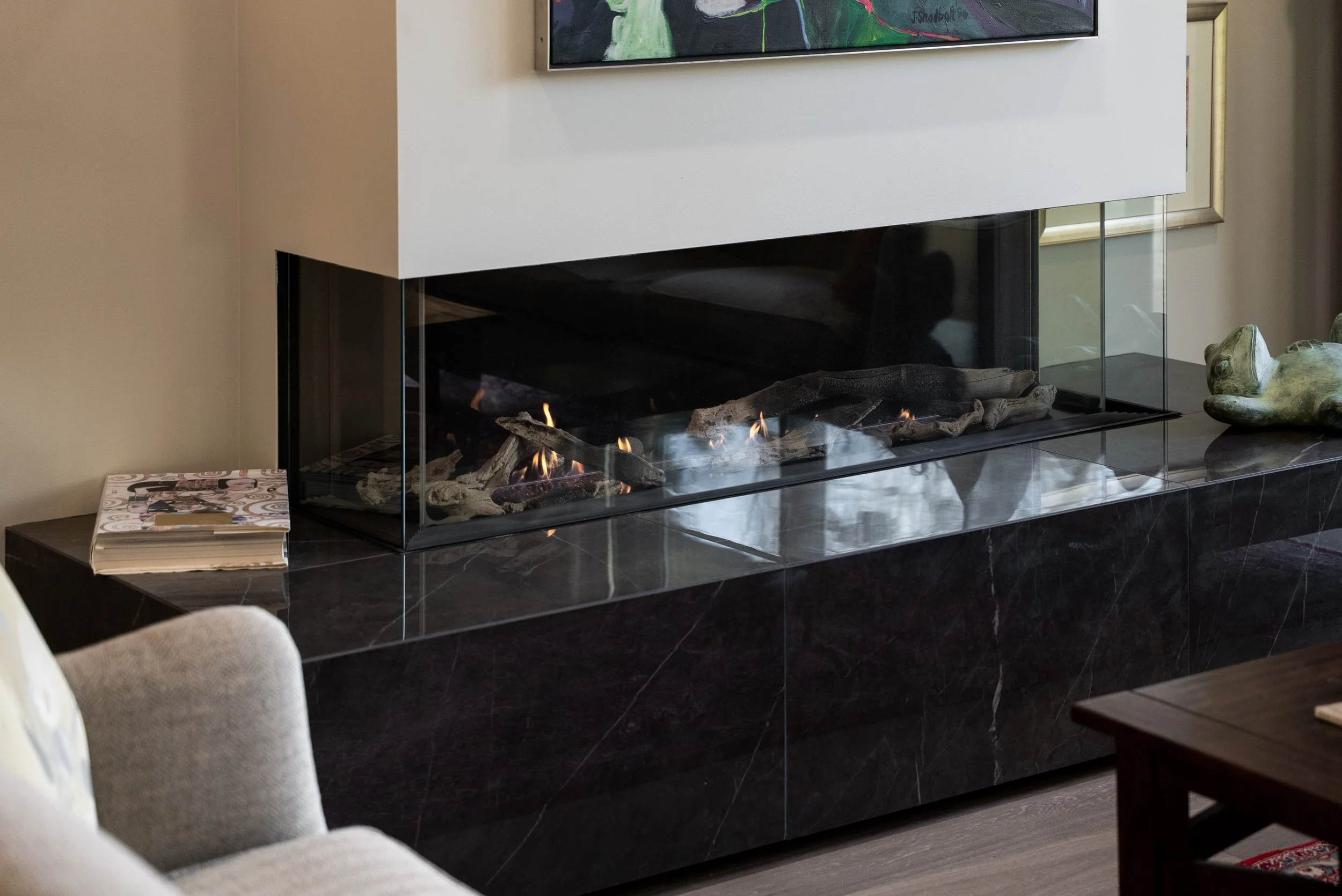 Modern living room fireplace with glass enclosure, black marble surround, and decorative items including a green sculpture and a stack of magazines.