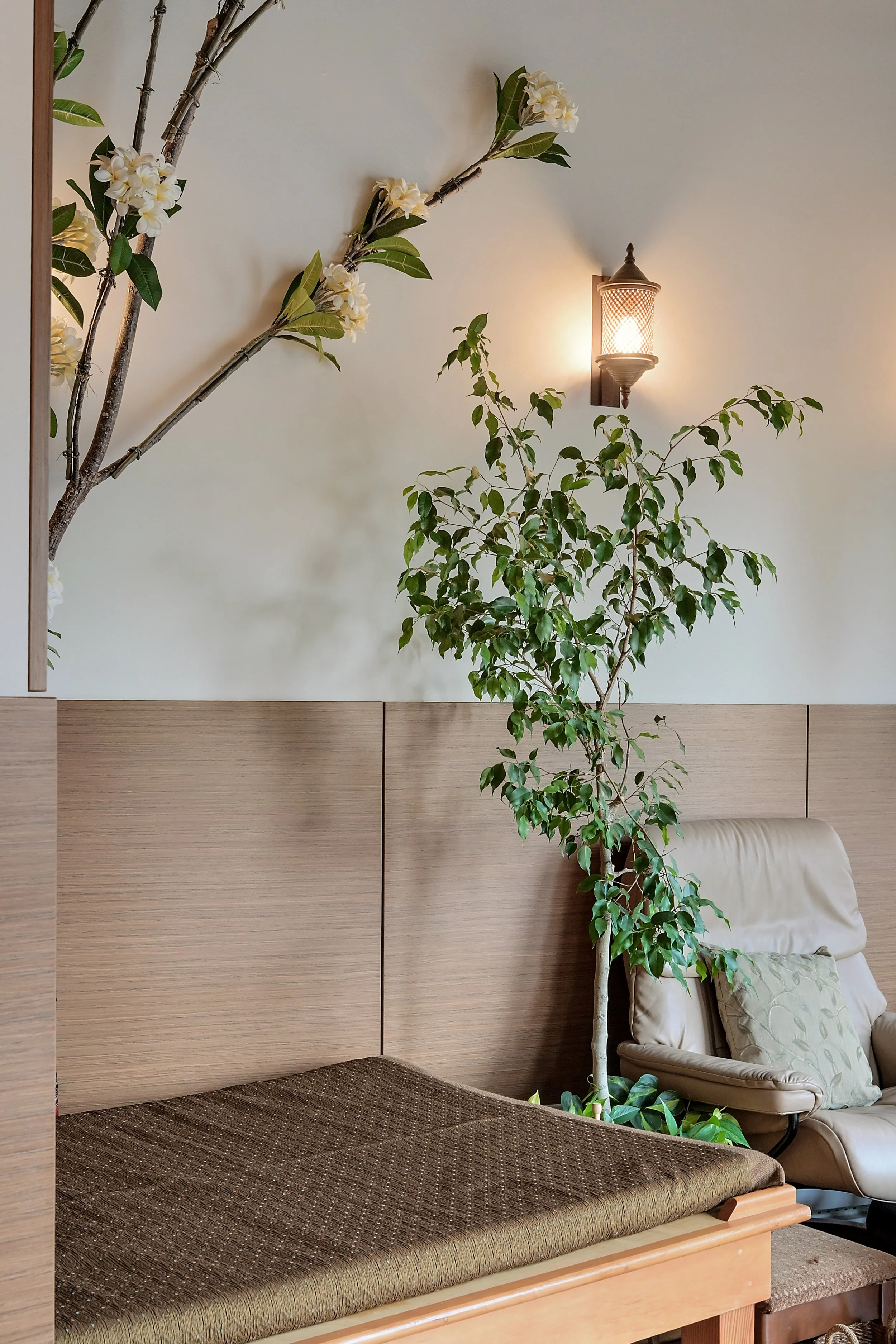 A cozy indoor corner with a wooden daybed with a cushioned top, a beige armchair with a pillow, a tall green plant, a wall-mounted light fixture, and a branch with white flowers and green leaves extending across the wall.