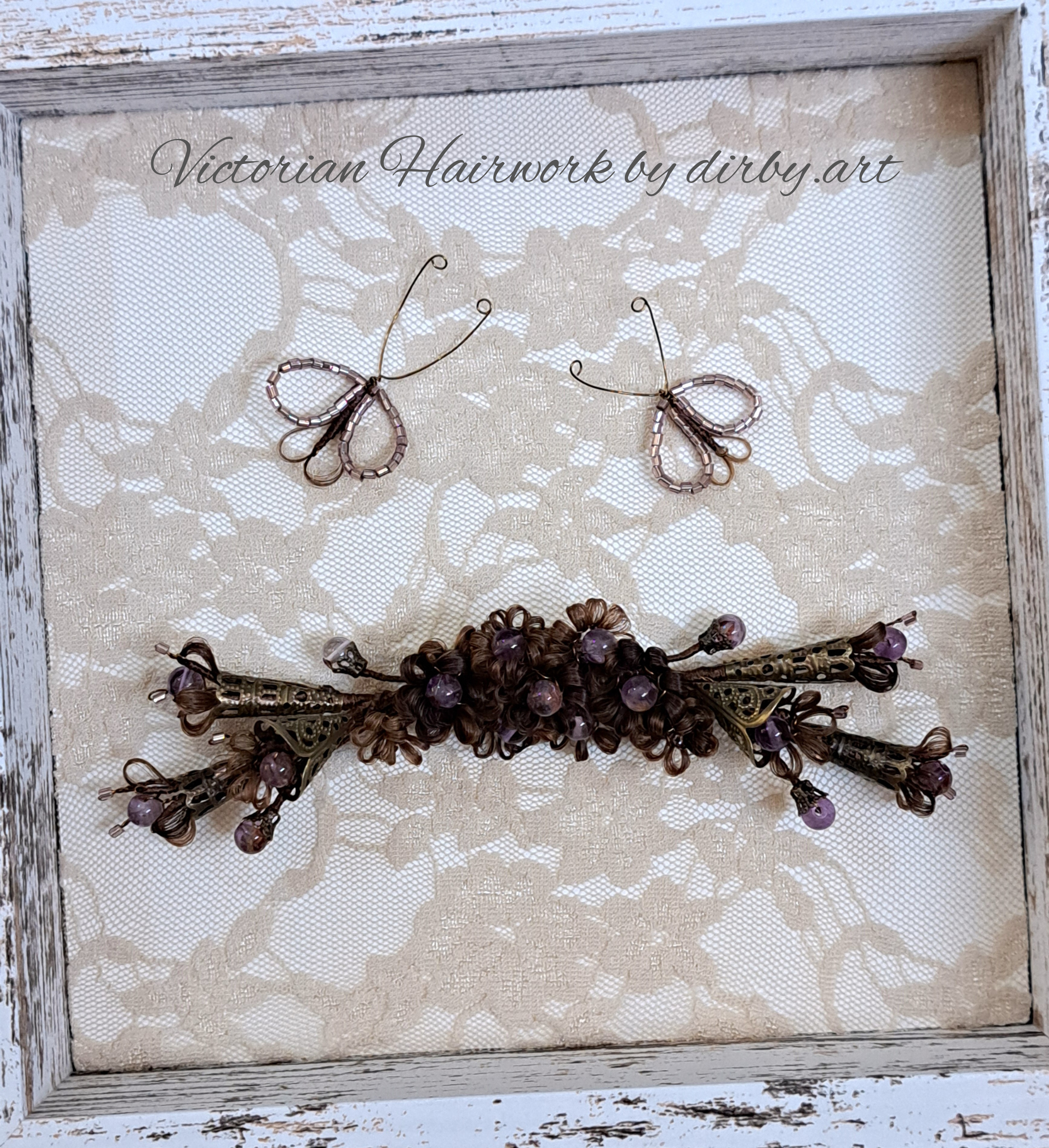 A Victorian-style hair wreath made of human hair and purple beads, with intricate metalwork, displayed in a wooden frame with a lace background, created by historian and artist Diane Irby.