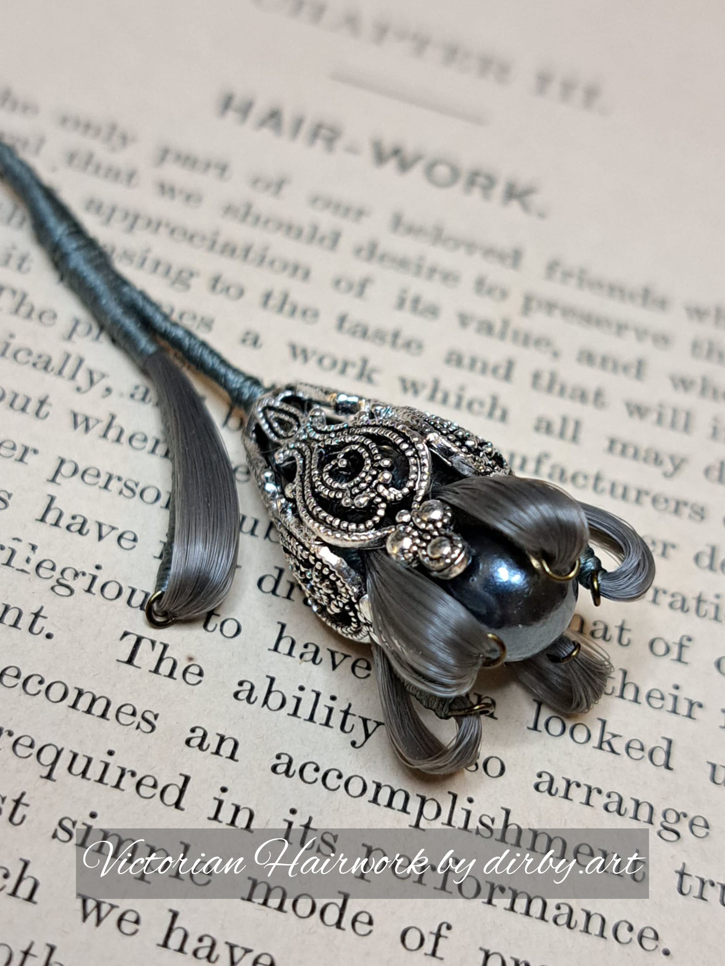 A flower made of human hair in a shade of grey with silver filigree embellishment, placed on an open antique book about Victorian hairwork, created by historian and artist Diane Irby.