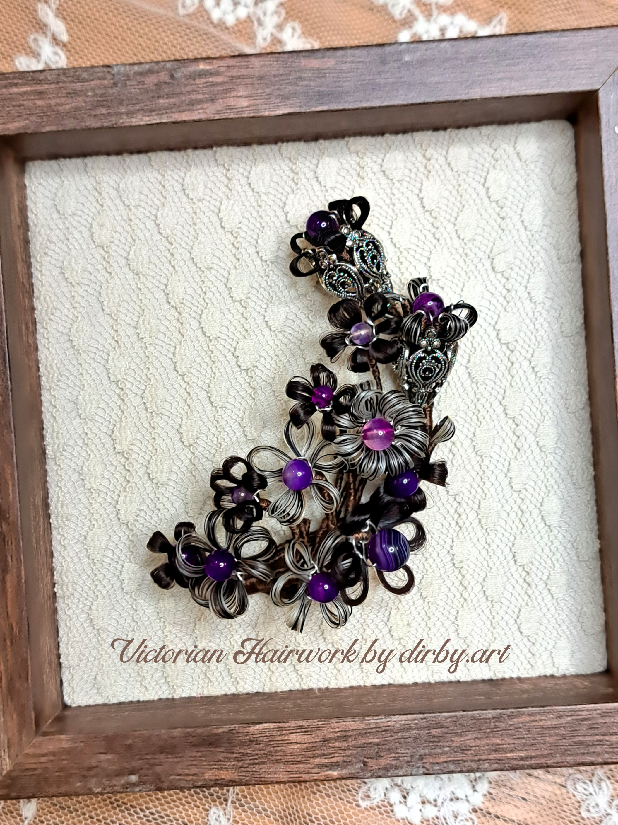 A Victorian-style hair wreath made of human and horse hair and purple beads, with intricate metalwork, displayed in a wooden frame with a lace background, created by historian and artist Diane Irby.