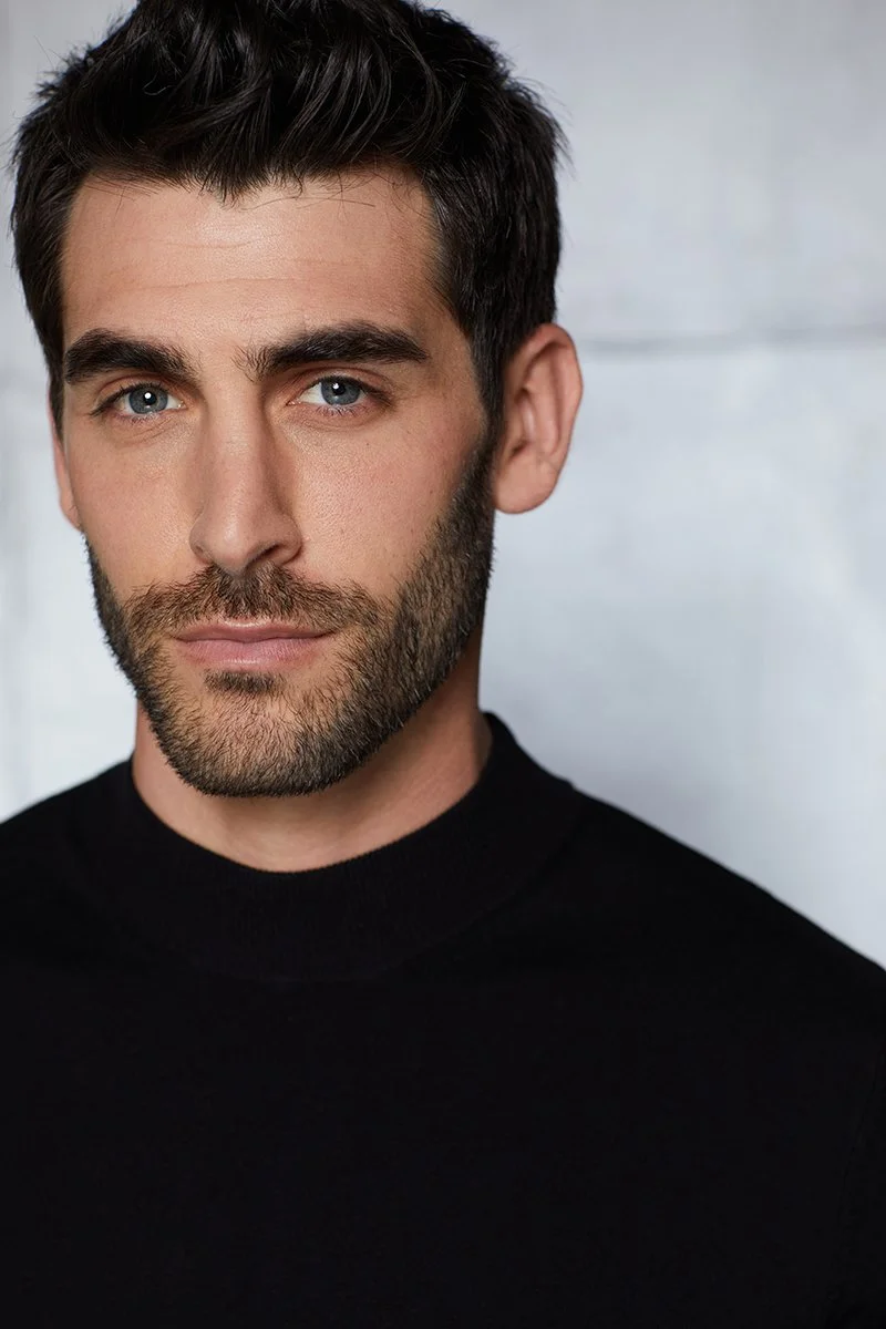 Close-up of a man's face with blue eyes, dark hair, and facial hair, wearing a black shirt, against a light background.